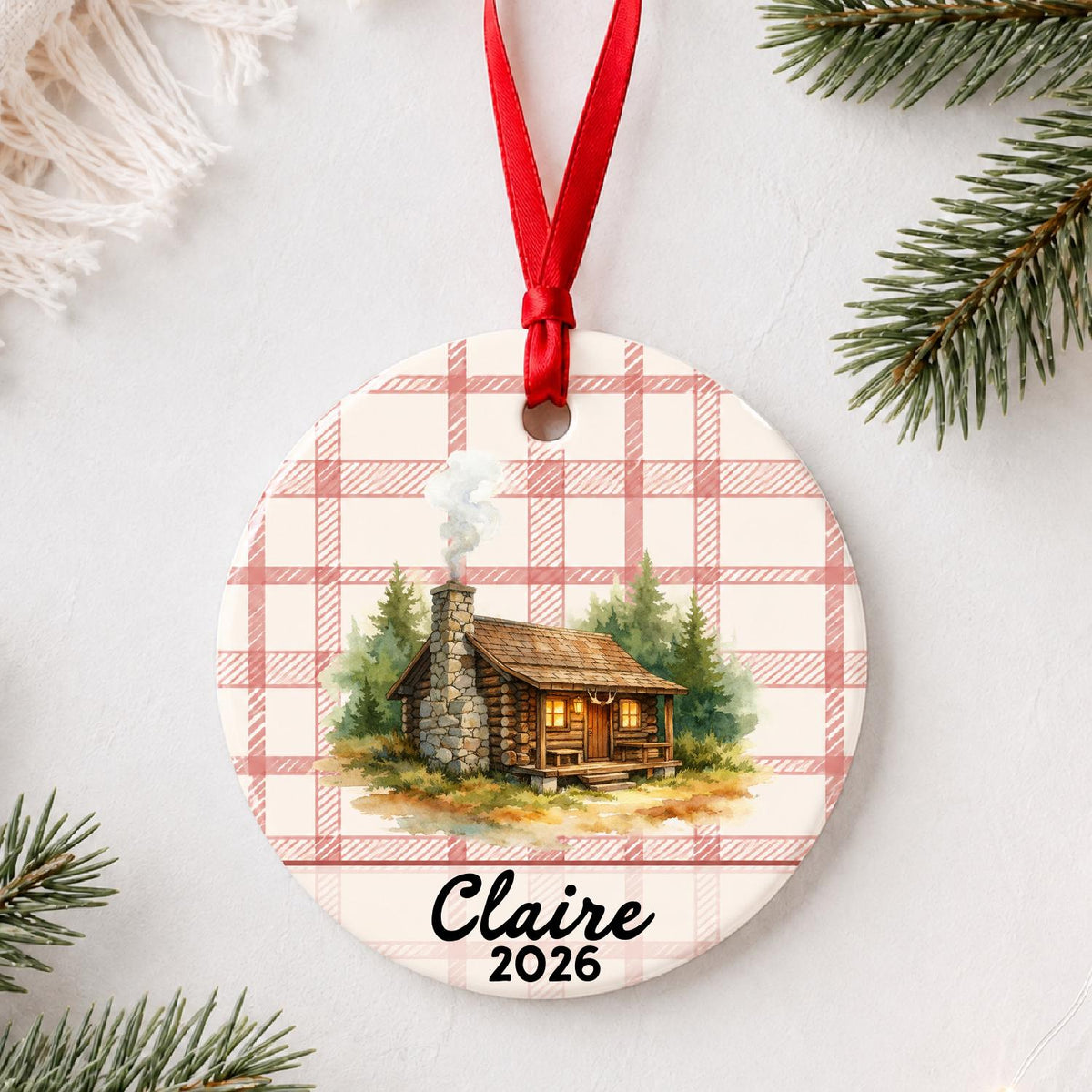 Personalized log cabin ceramic ornament with name and year on red plaid styled with pine branches on a soft white background