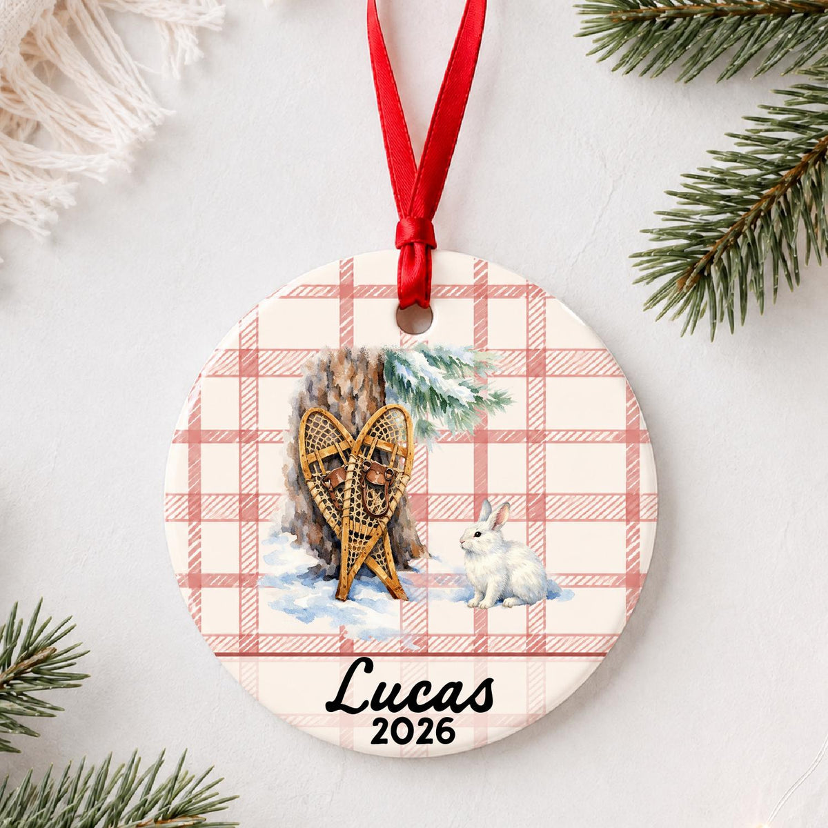 Personalized snowshoes and snowshoe hare ceramic ornament with name and year on red plaid styled with pine branches on a soft white background