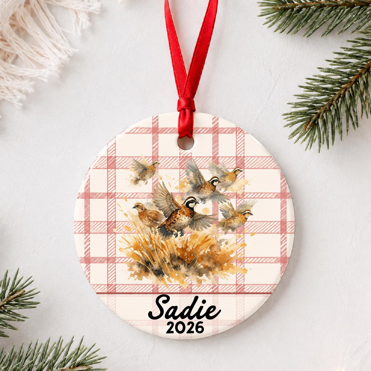 Personalized bobwhite quail ceramic ornament with name and year on red plaid styled with pine branches on a soft white background