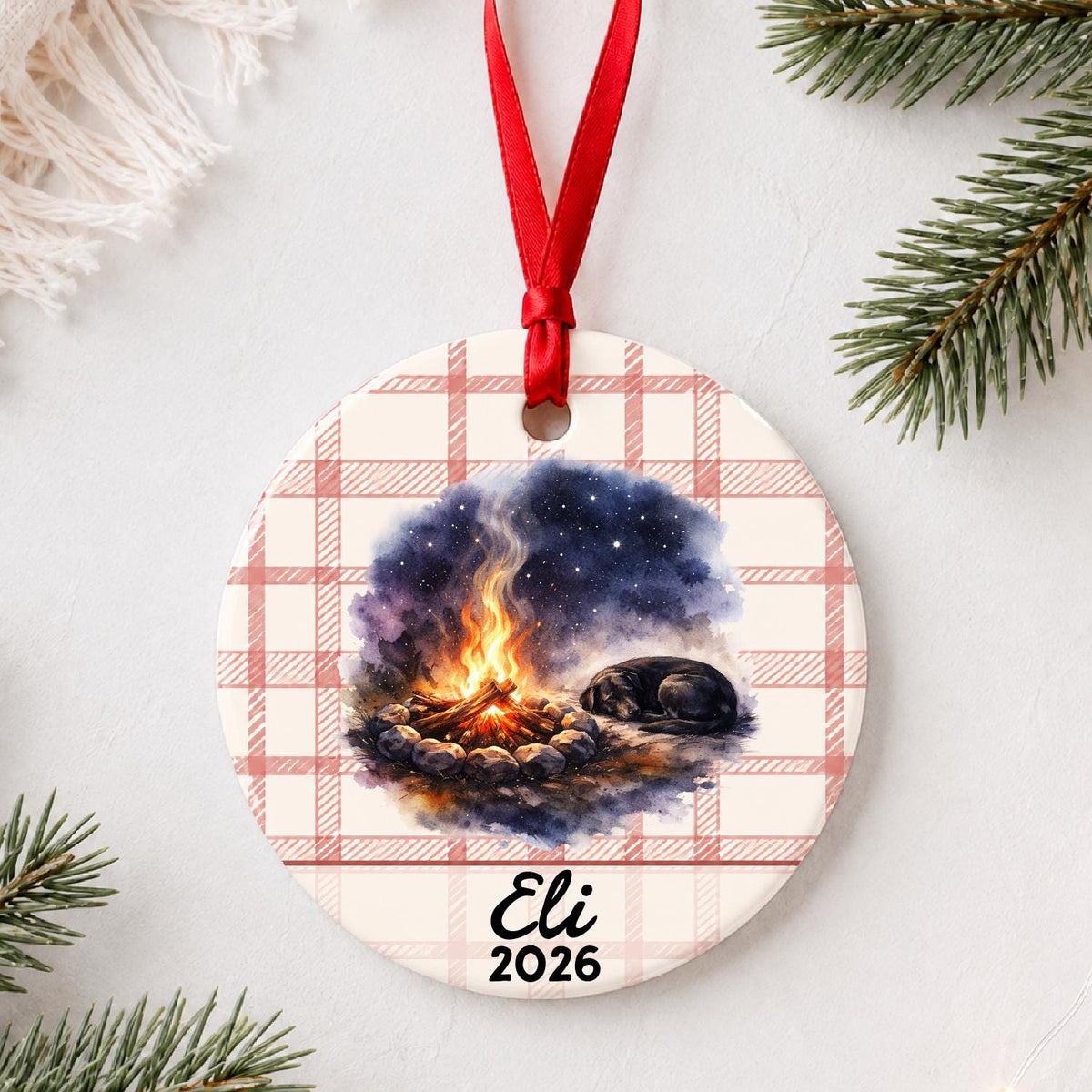 Personalized campfire and dog ceramic ornament with name and year on red plaid styled with pine branches on a soft white background