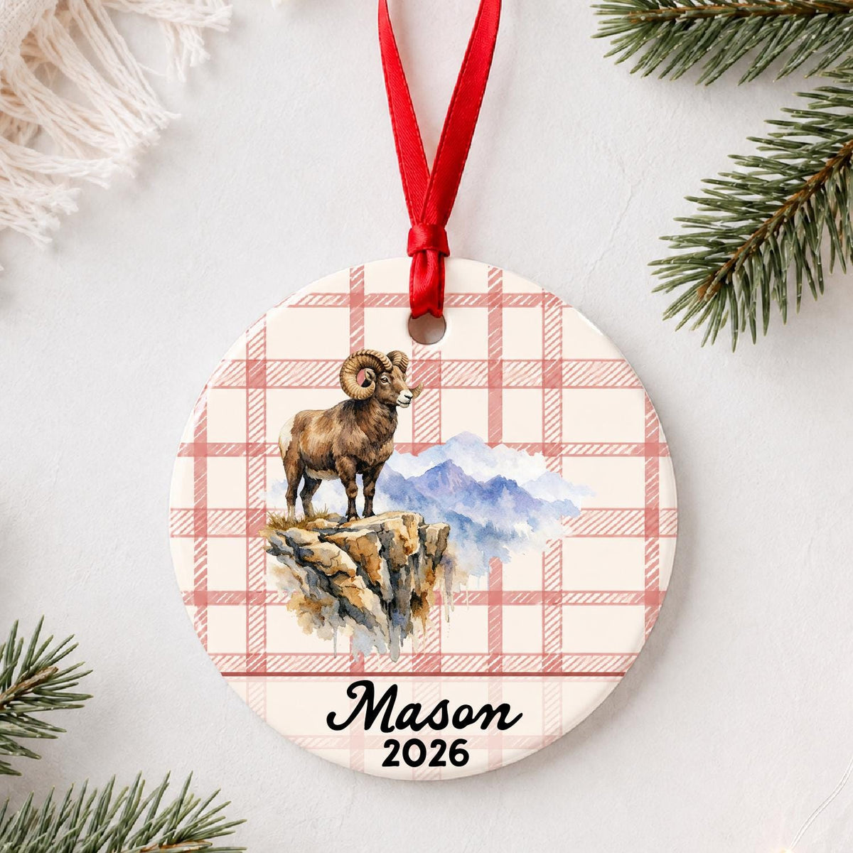 Personalized bighorn sheep ceramic ornament with name and year on red plaid styled with pine branches on a soft white background
