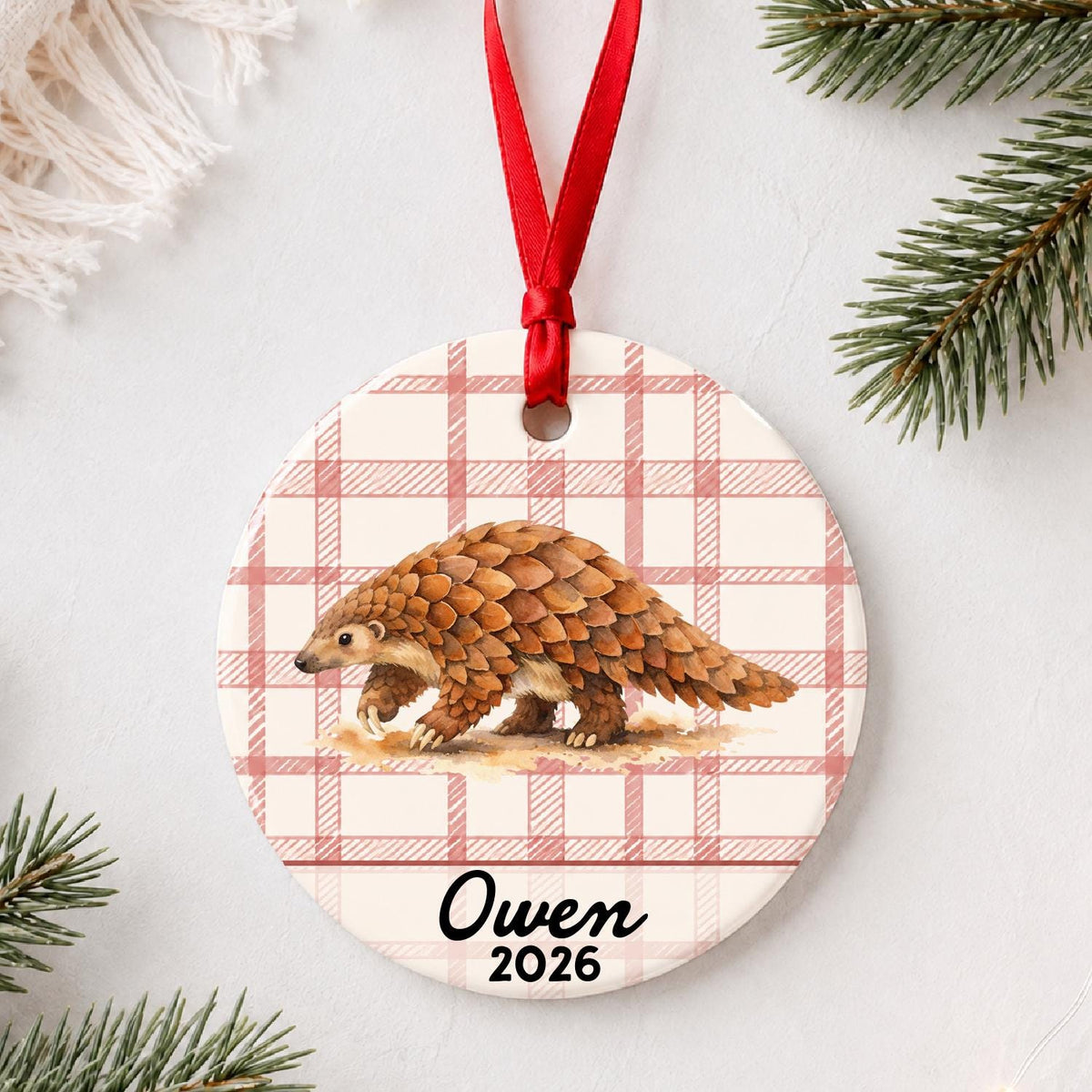 Personalized pangolin ceramic ornament with name and year on red plaid styled with pine branches on a soft white background
