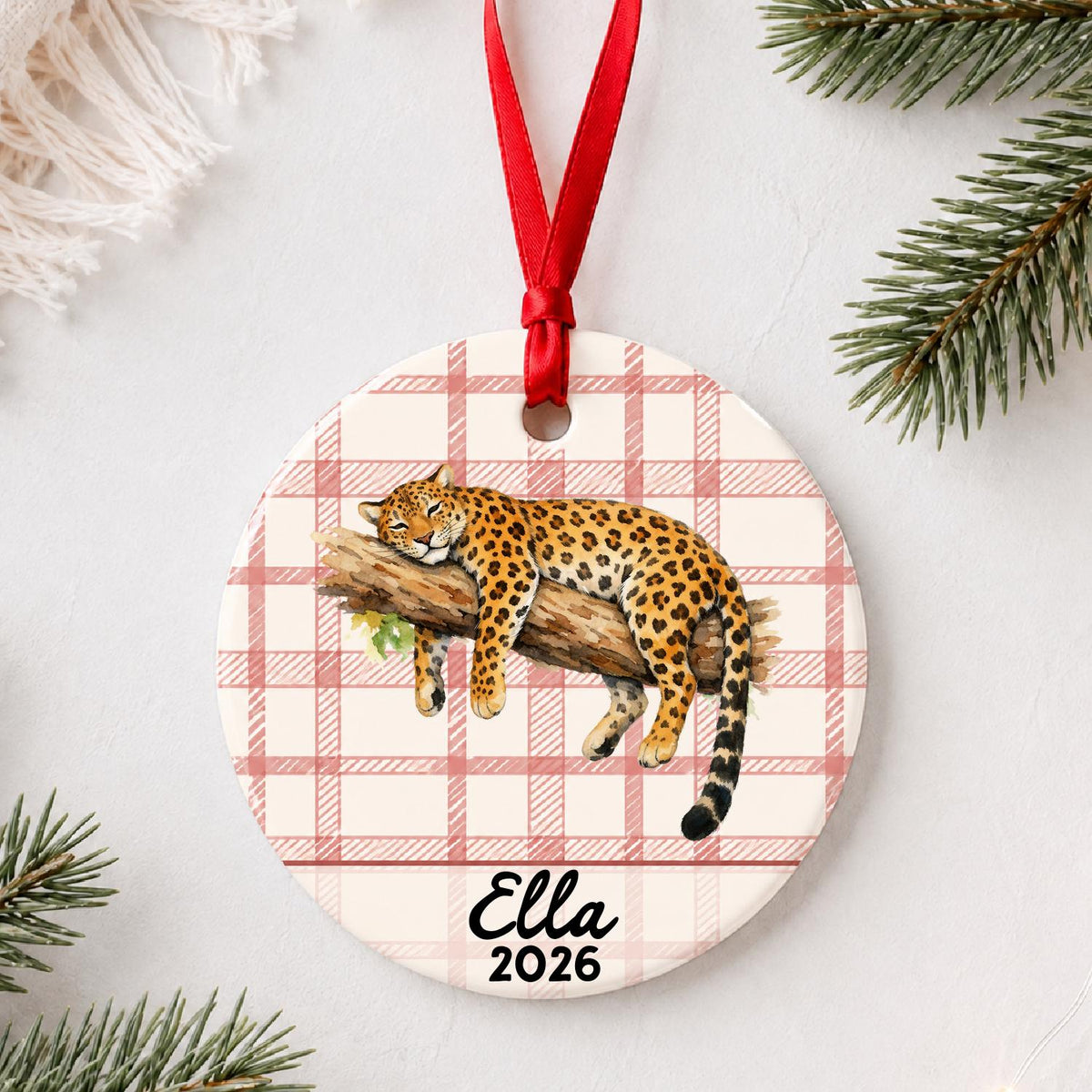 Personalized leopard ceramic ornament with name and year on red plaid styled with pine branches on a soft white background