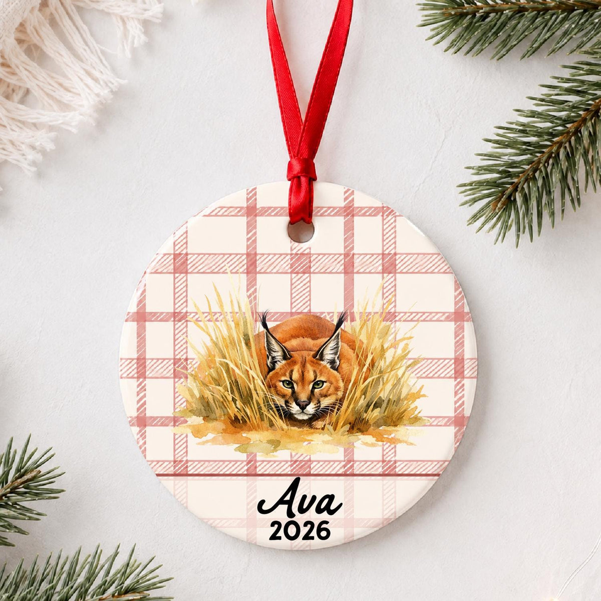 Personalized caracal ceramic ornament with name and year on red plaid styled with pine branches on a soft white background