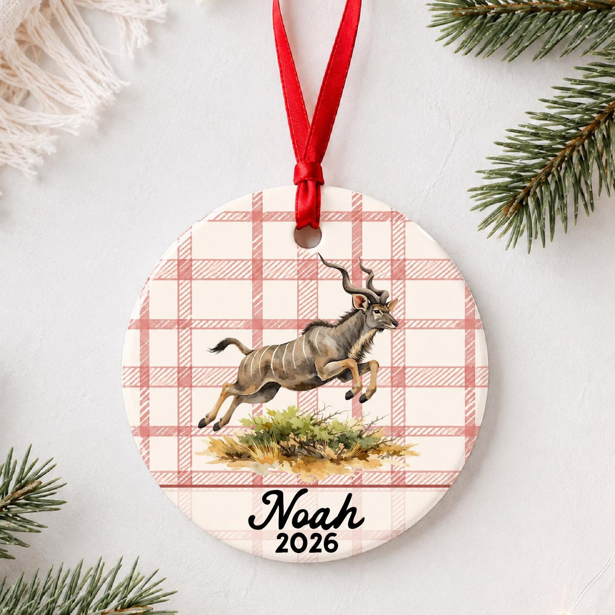 Personalized kudu ceramic ornament with name and year on red plaid styled with pine branches on a soft white background