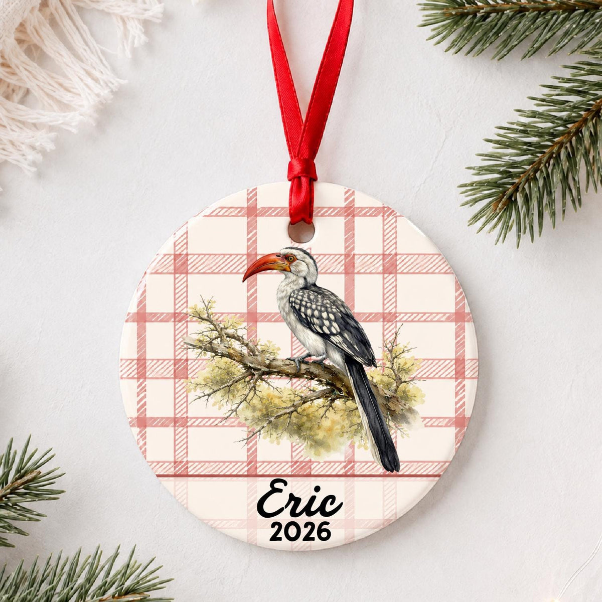 Personalized hornbill ceramic ornament with name and year on red plaid styled with pine branches on a soft white background