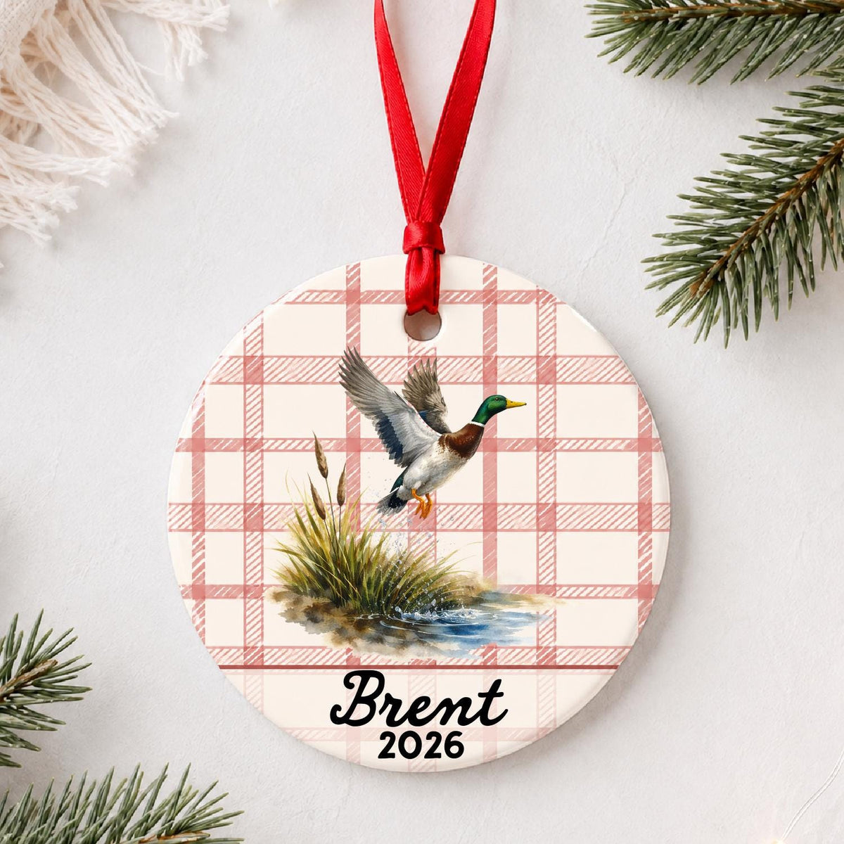 Personalized mallard duck ceramic ornament with name and year on red plaid styled with pine branches on a soft white background