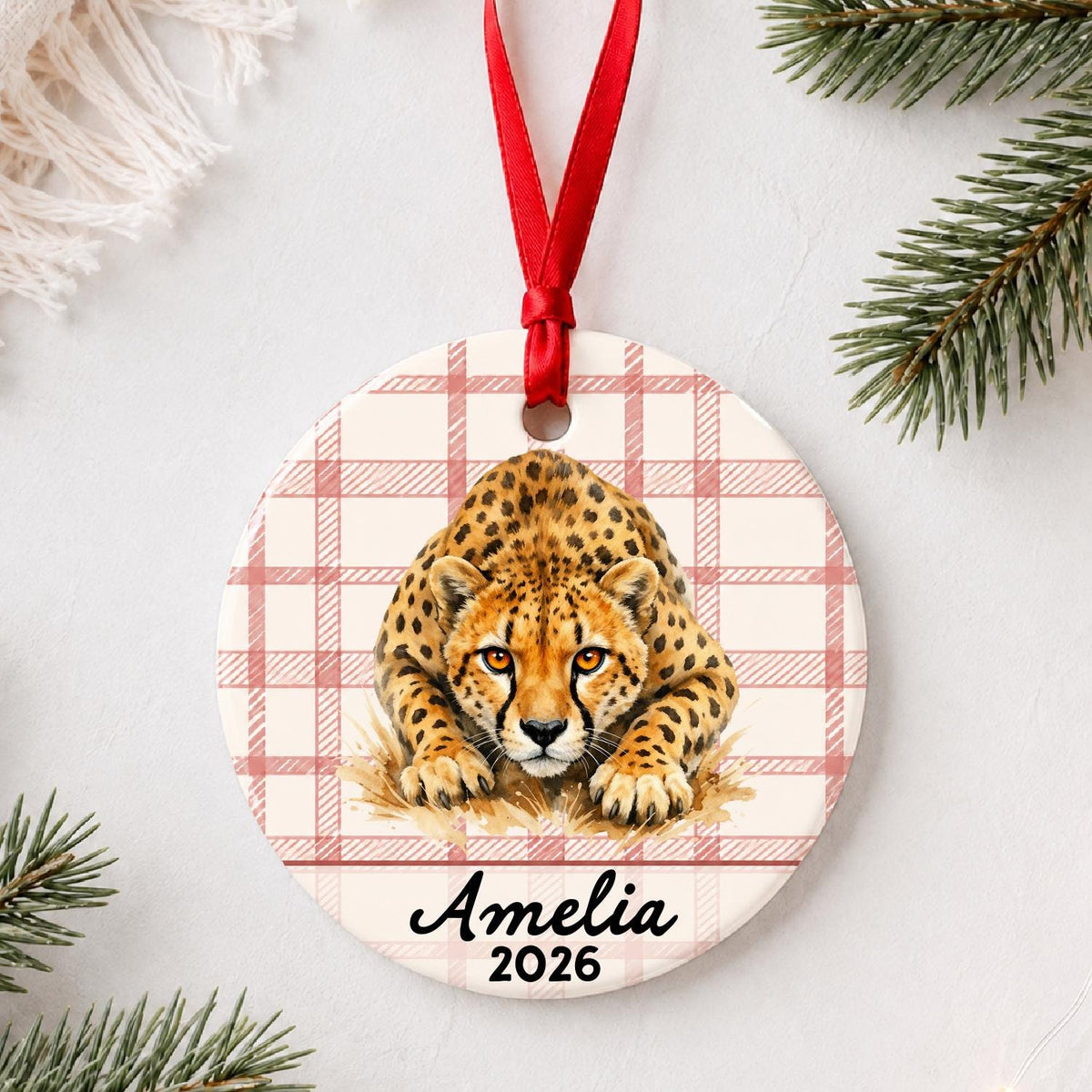 Personalized cheetah ceramic ornament with name and year on red plaid styled with pine branches on a soft white background