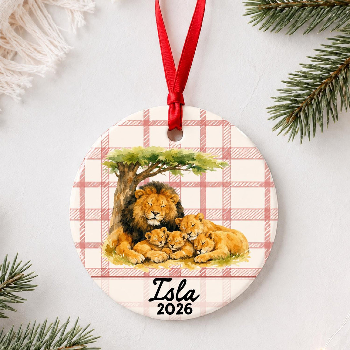 Personalized lion pride ceramic ornament with name and year on red plaid styled with pine branches on a soft white background