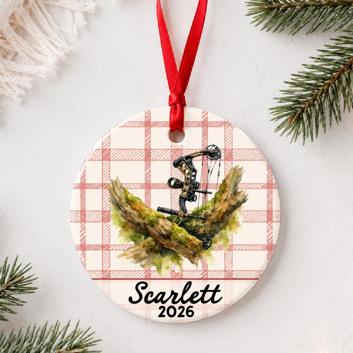 Personalized compound bow ceramic ornament with name and year on red plaid styled with pine branches on a soft white background