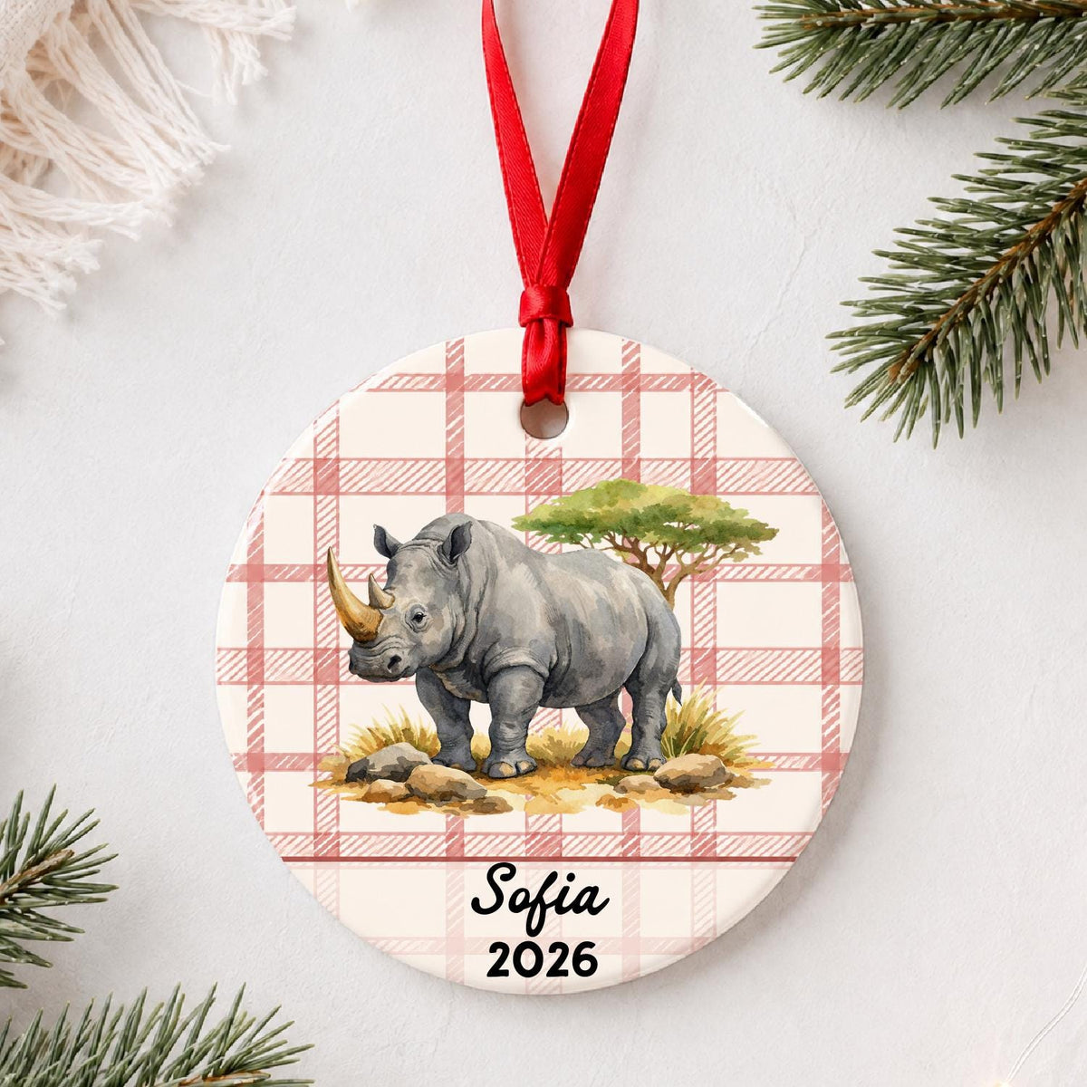 Personalized white rhinoceros ceramic ornament with name and year on red plaid styled with pine branches on a soft white background
