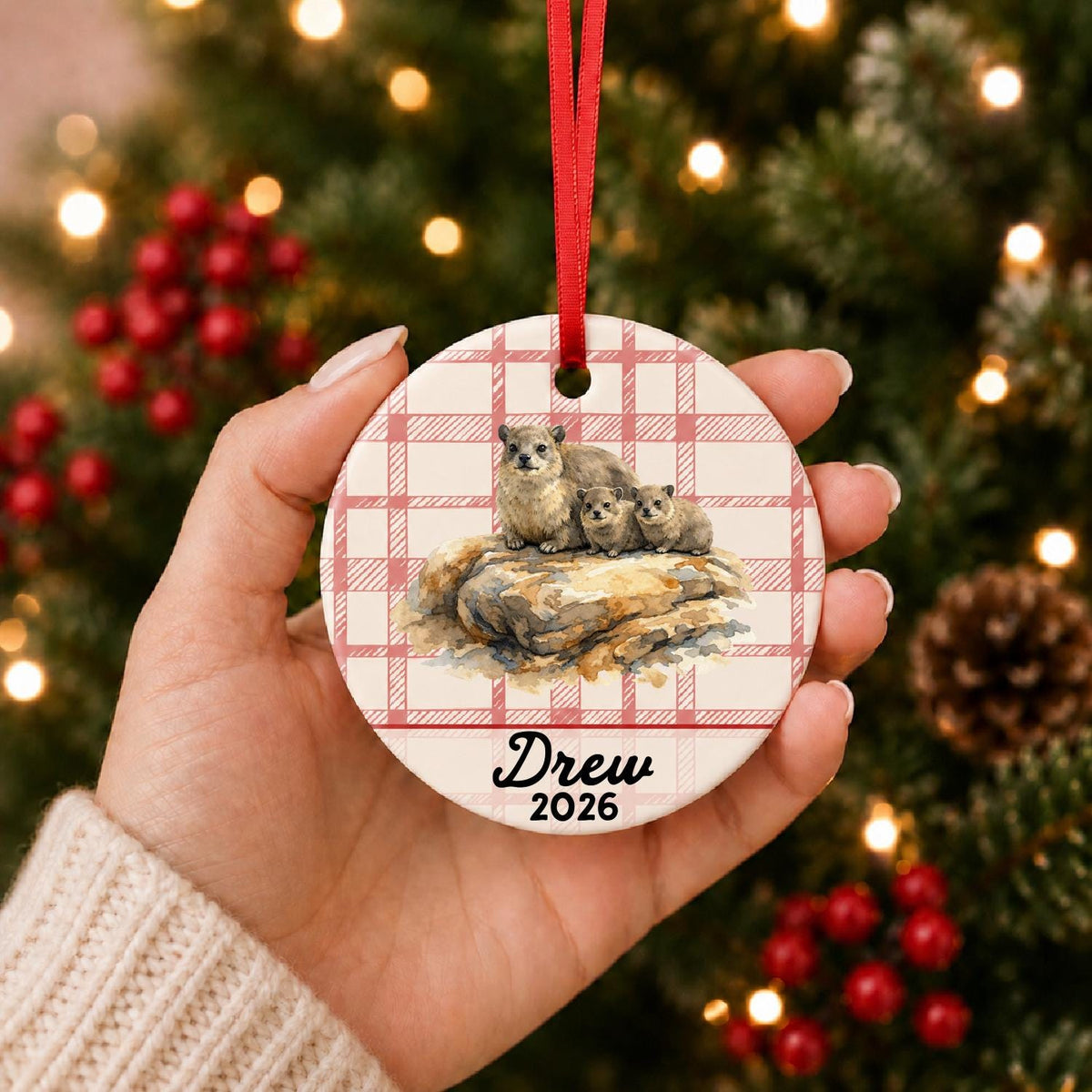 Hand holding the personalized rock hyraxes ceramic ornament with name and year on red plaid in front of a Christmas tree