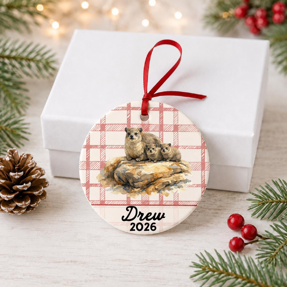 Personalized rock hyraxes ceramic ornament with name and year on red plaid in front of a white gift box with pinecones and red berries