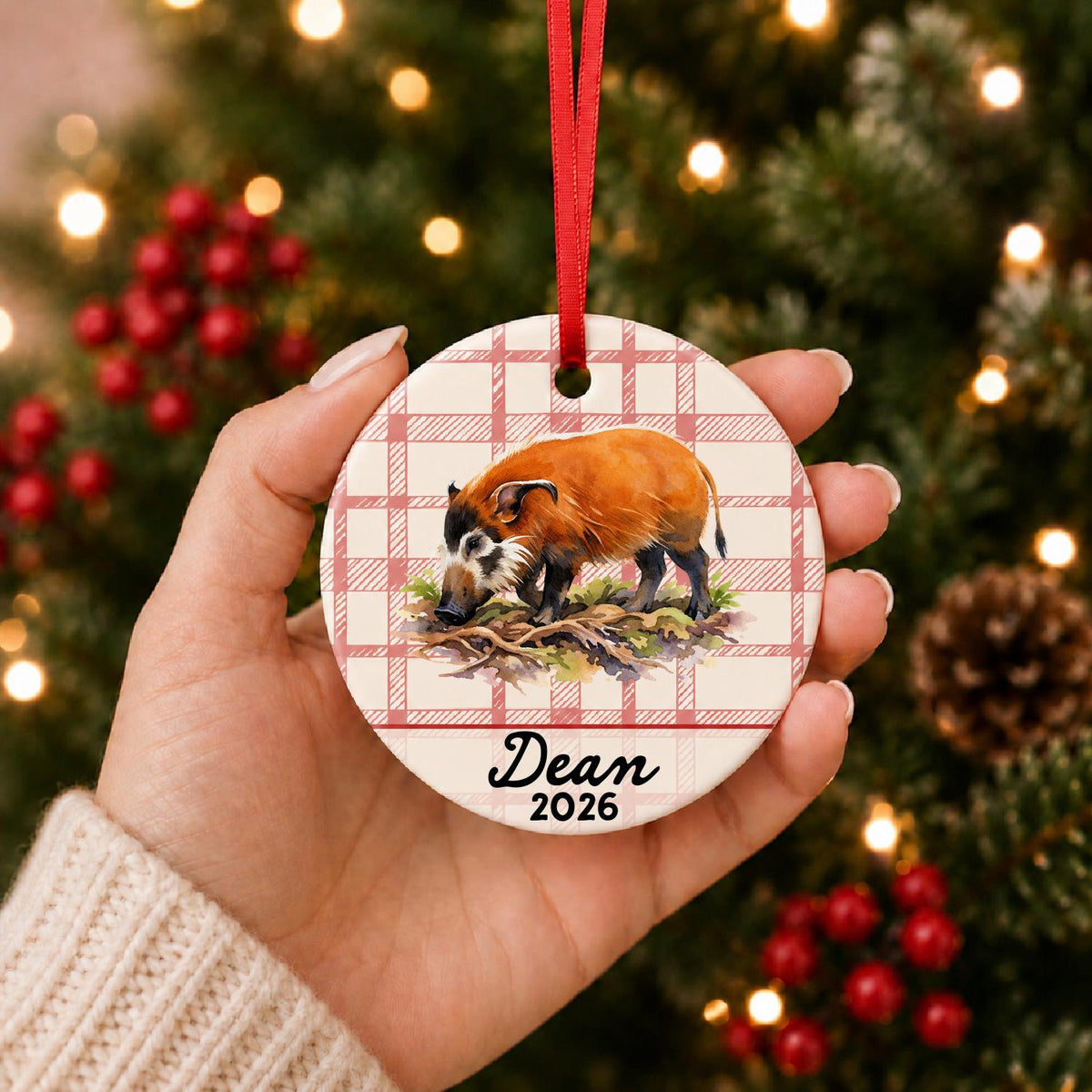 Hand holding the personalized red river hog ceramic ornament with name and year on red plaid in front of a Christmas tree