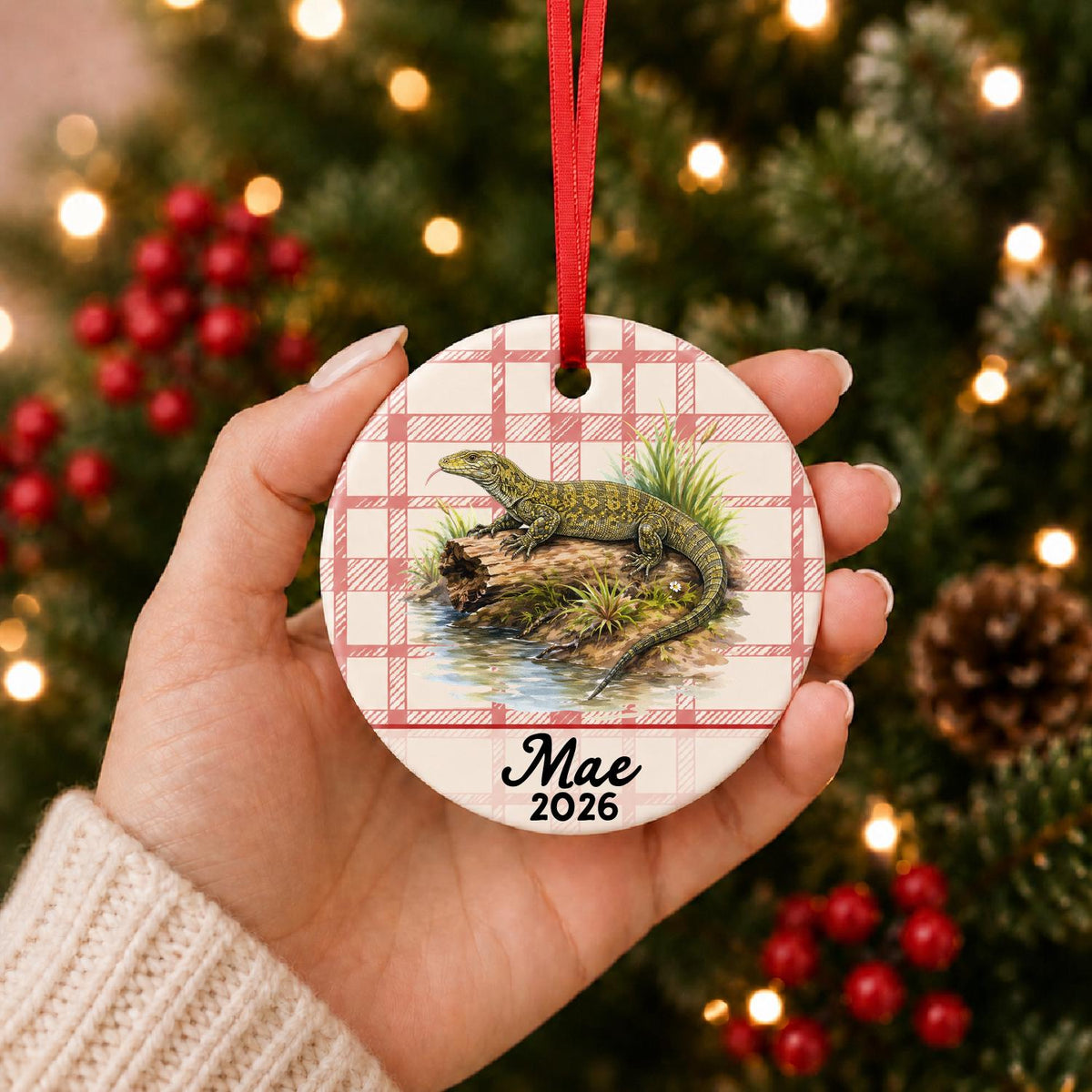 Hand holding the personalized monitor lizard ceramic ornament with name and year on red plaid in front of a Christmas tree