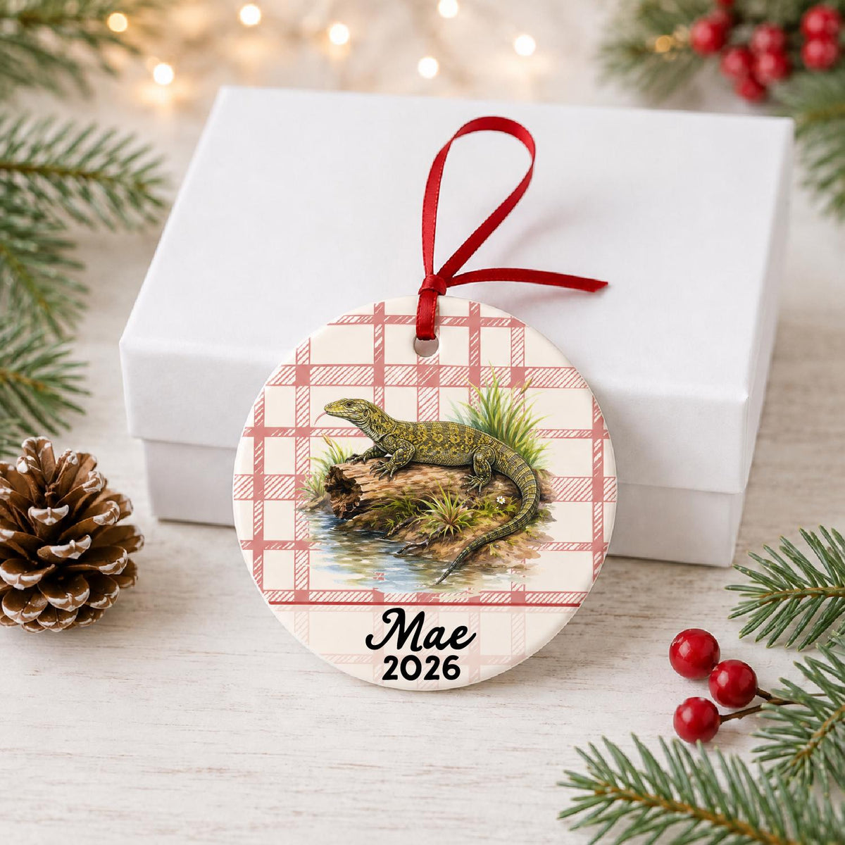 Personalized monitor lizard ceramic ornament with name and year on red plaid in front of a white gift box with pinecones and red berries