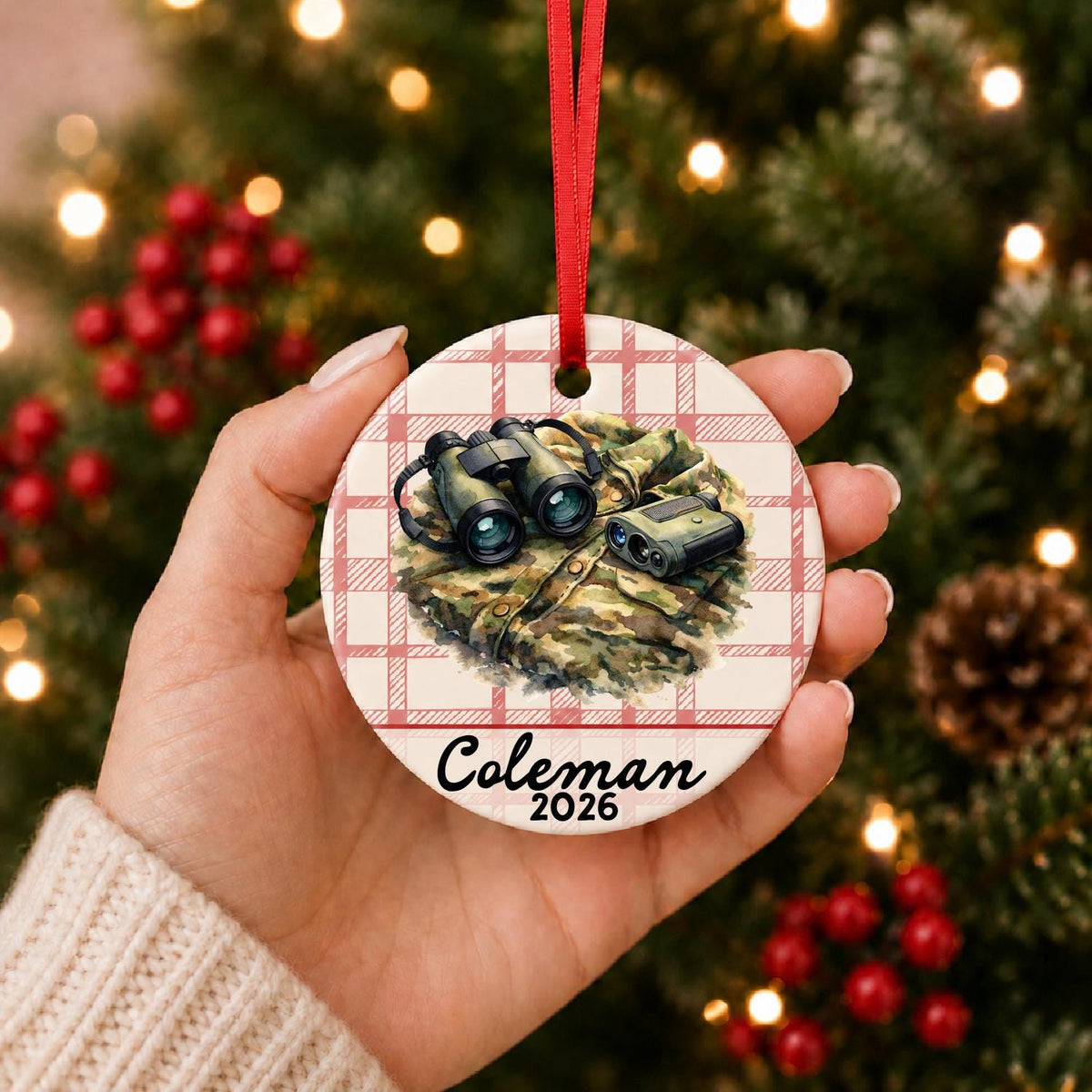Hand holding the personalized binoculars and rangefinder ceramic ornament with name and year on red plaid in front of a Christmas tree