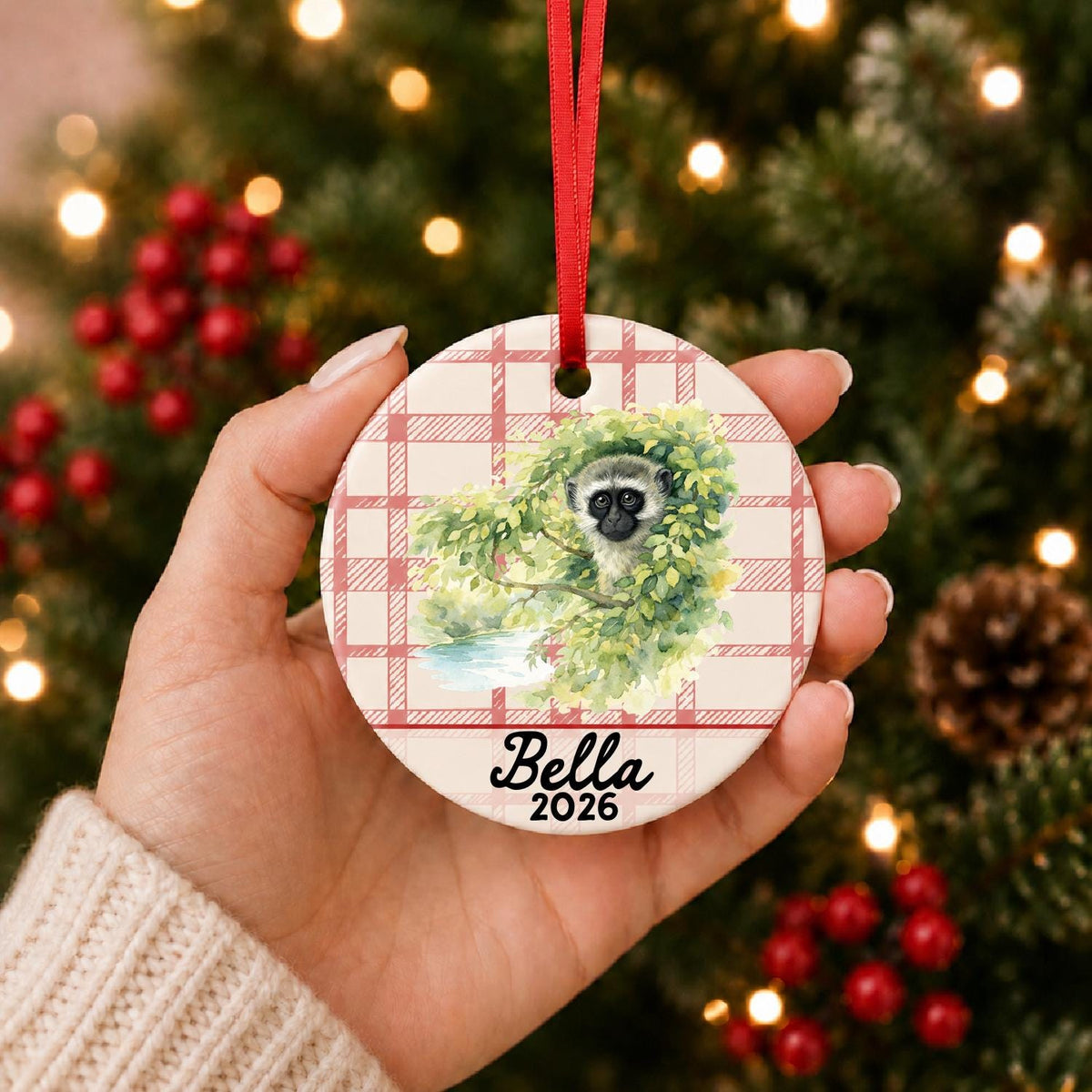 Hand holding the personalized vervet monkey ceramic ornament with name and year on red plaid in front of a Christmas tree