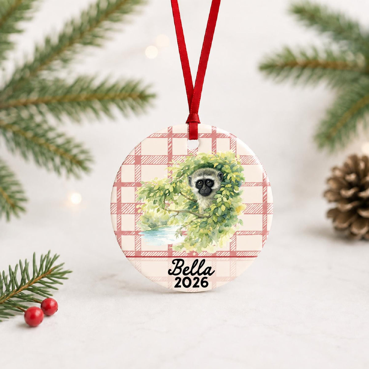 Personalized vervet monkey ceramic ornament with name and year on red plaid with red ribbon on a white surface with pine branches and pinecones