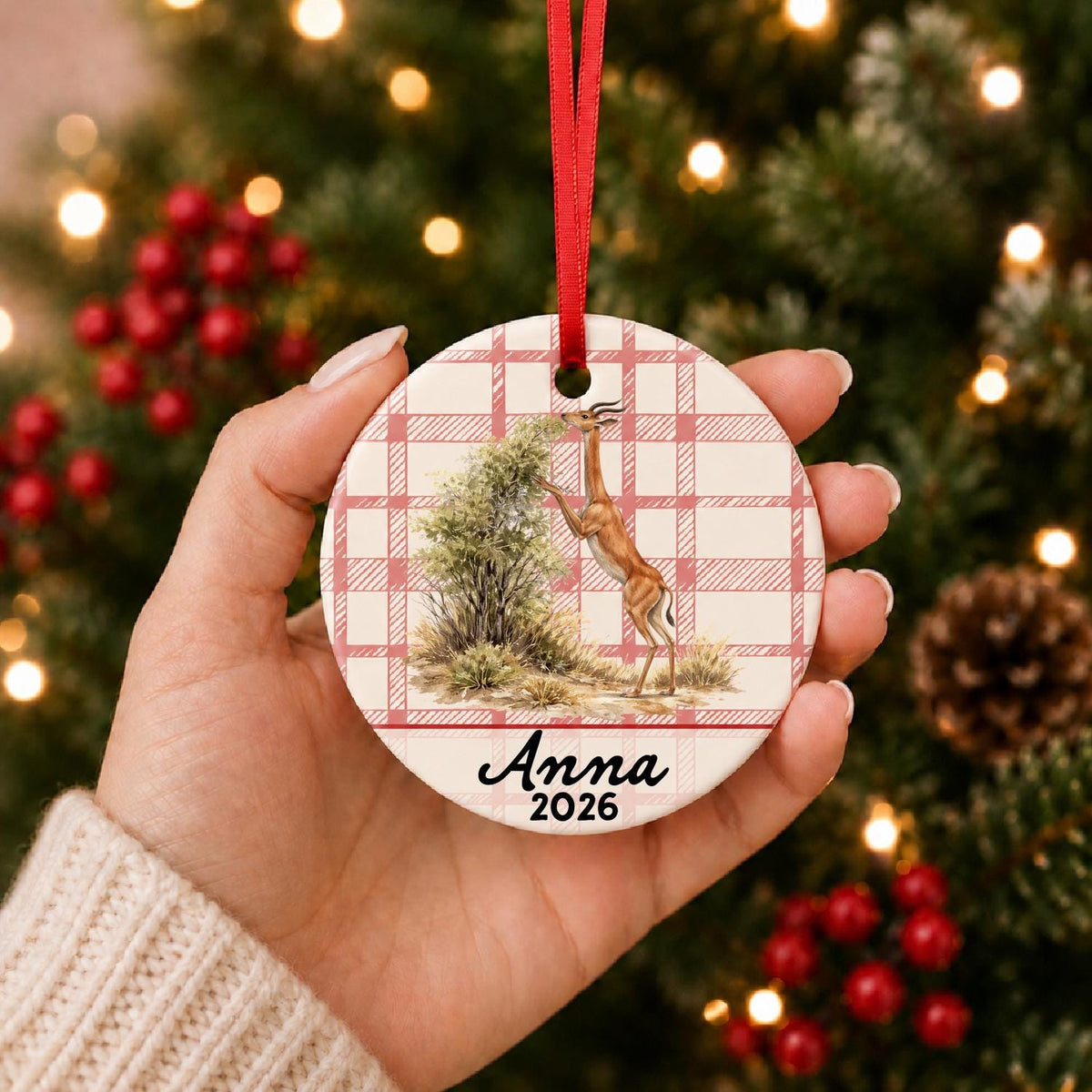 Hand holding the personalized gerenuk ceramic ornament with name and year on red plaid in front of a Christmas tree
