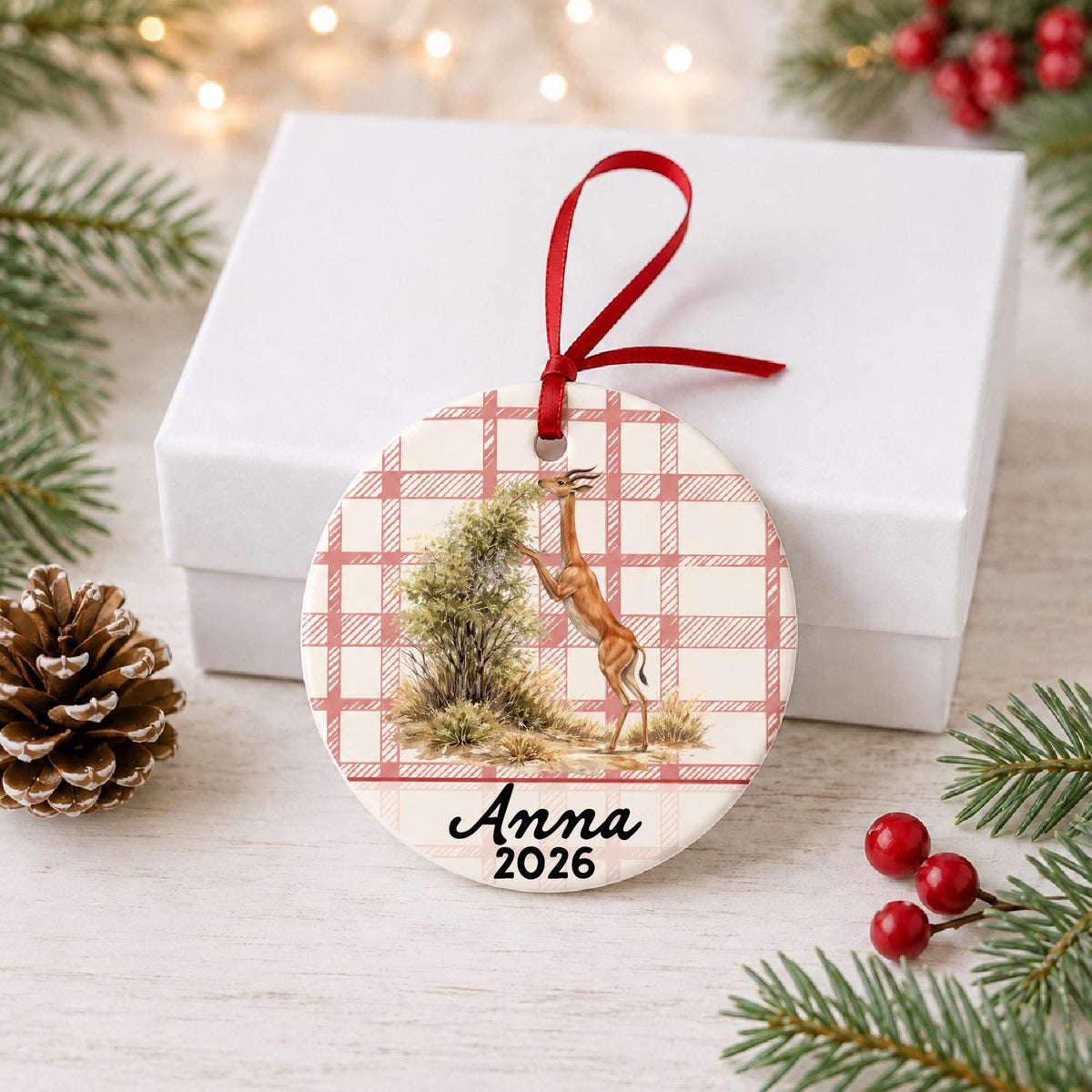 Personalized gerenuk ceramic ornament with name and year on red plaid in front of a white gift box with pinecones and red berries