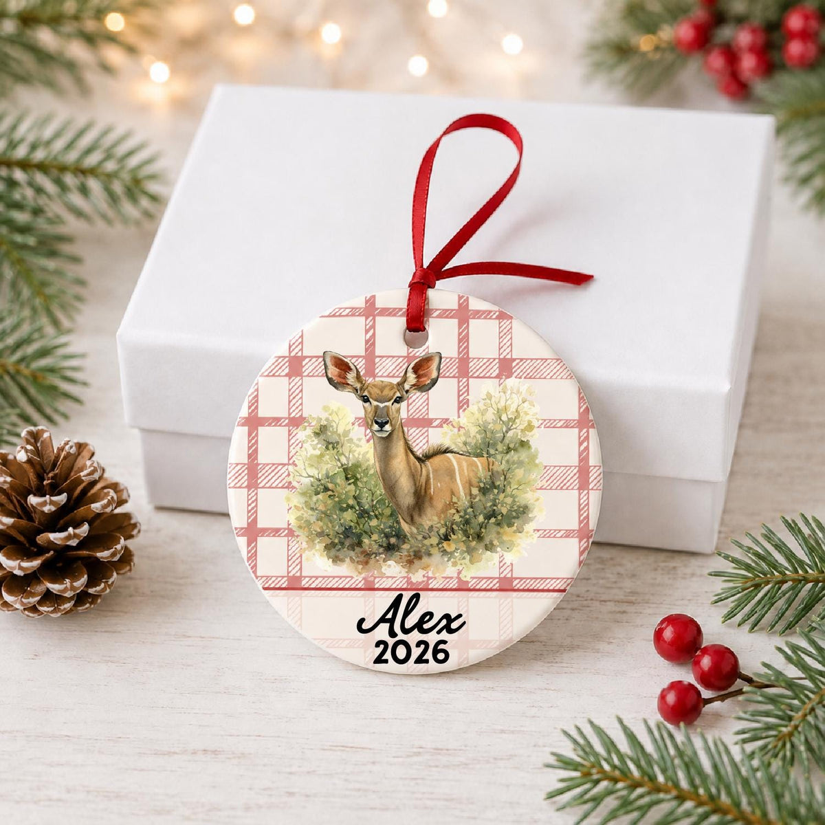 Personalized kudu in woodland ceramic ornament with name and year on red plaid in front of a white gift box with pinecones and red berries
