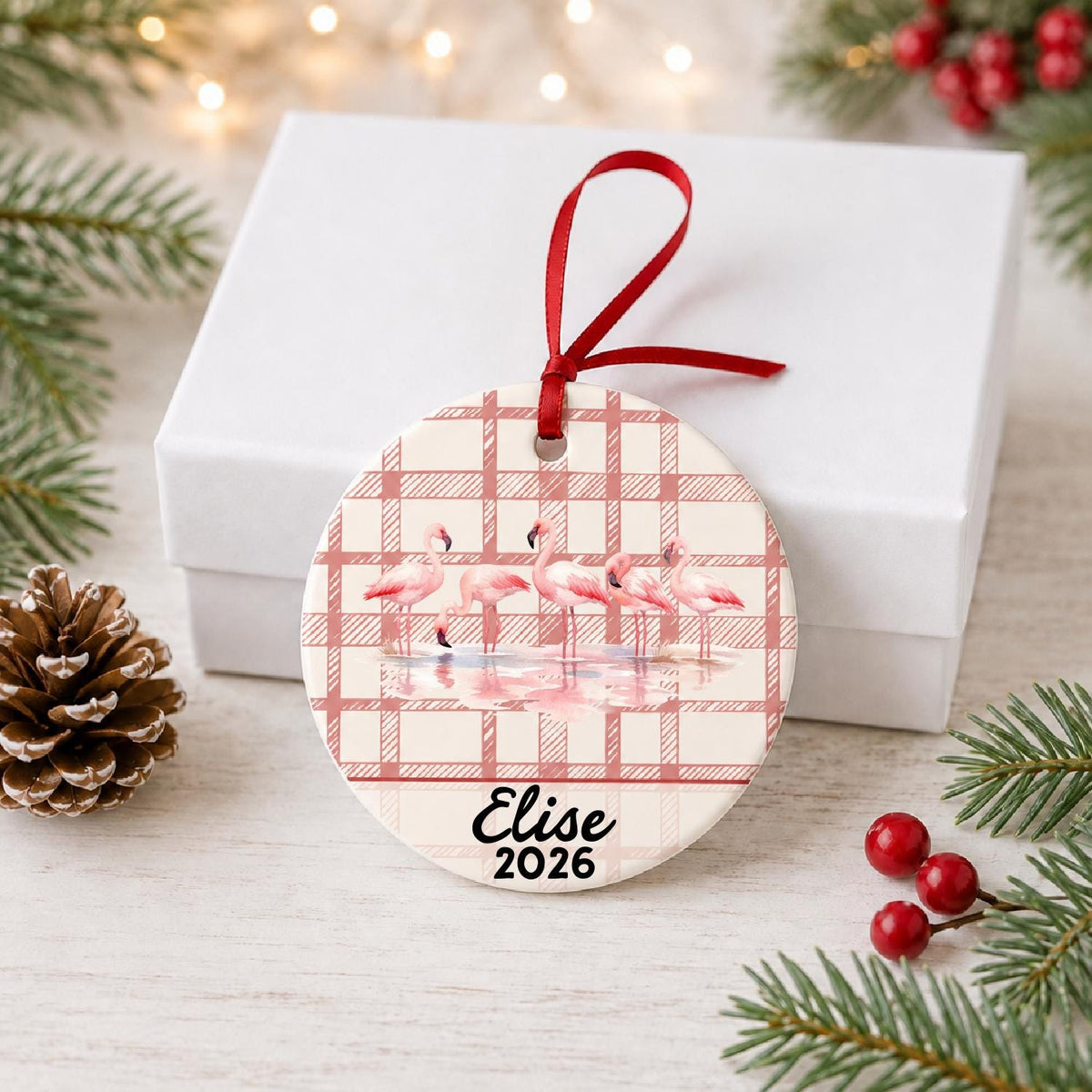 Personalized flamingo flock ceramic ornament with name and year on red plaid in front of a white gift box with pinecones and red berries