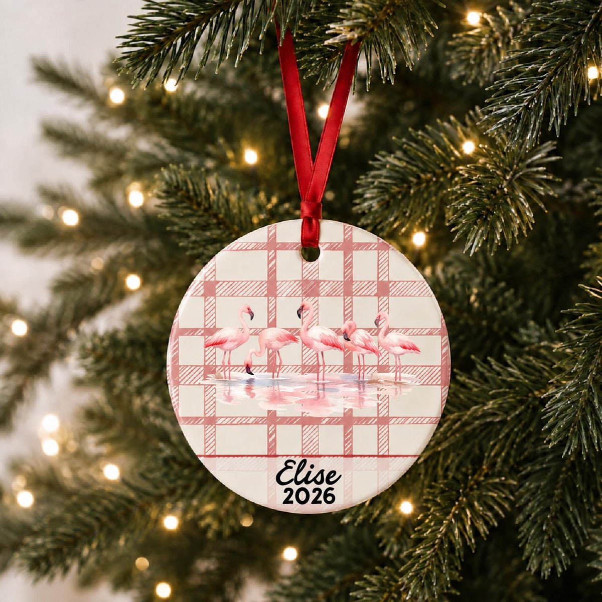 Personalized flamingo flock ceramic ornament with name and year on red plaid hanging on a lit Christmas tree
