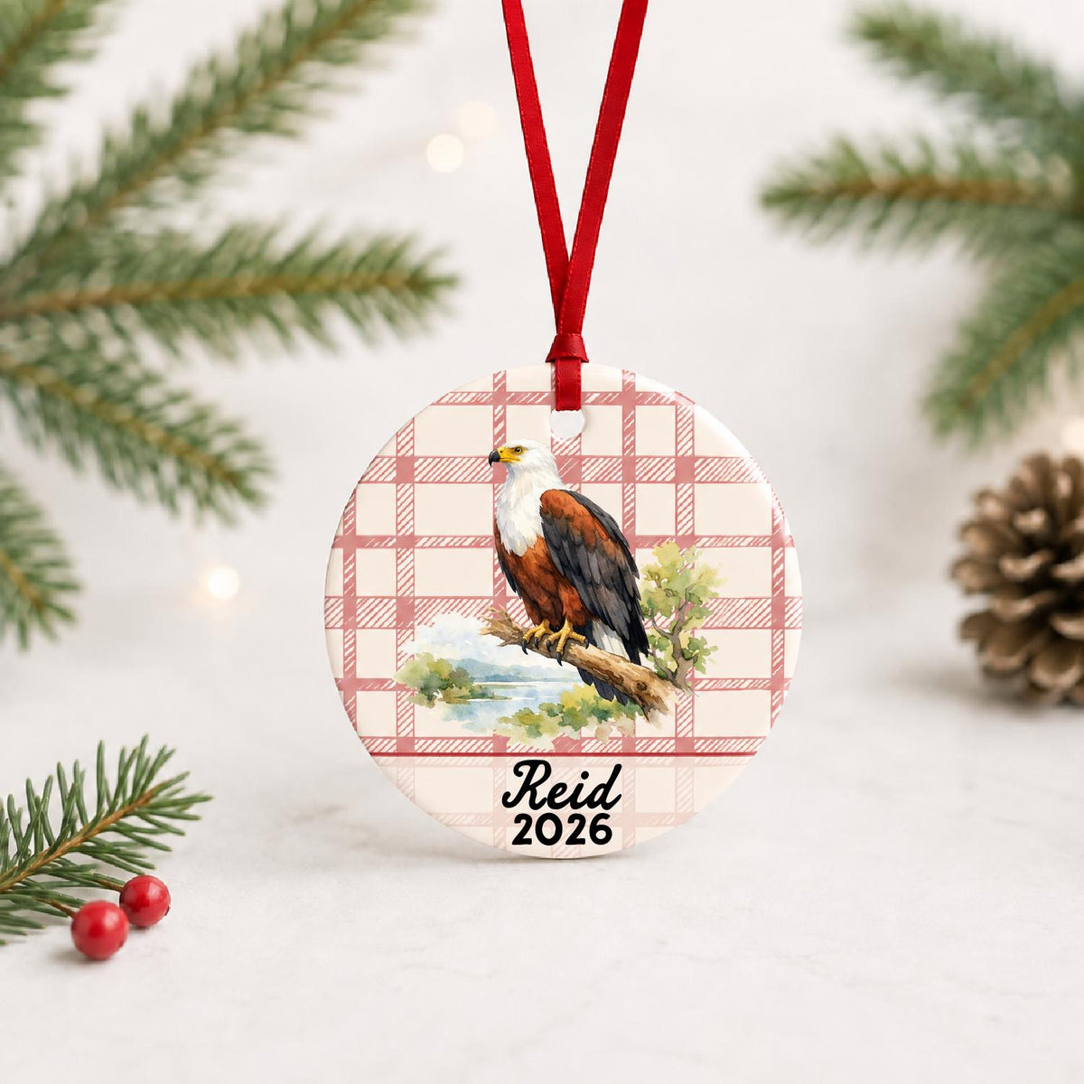 Personalized african fish eagle ceramic ornament with name and year on red plaid with red ribbon on a white surface with pine branches and pinecones