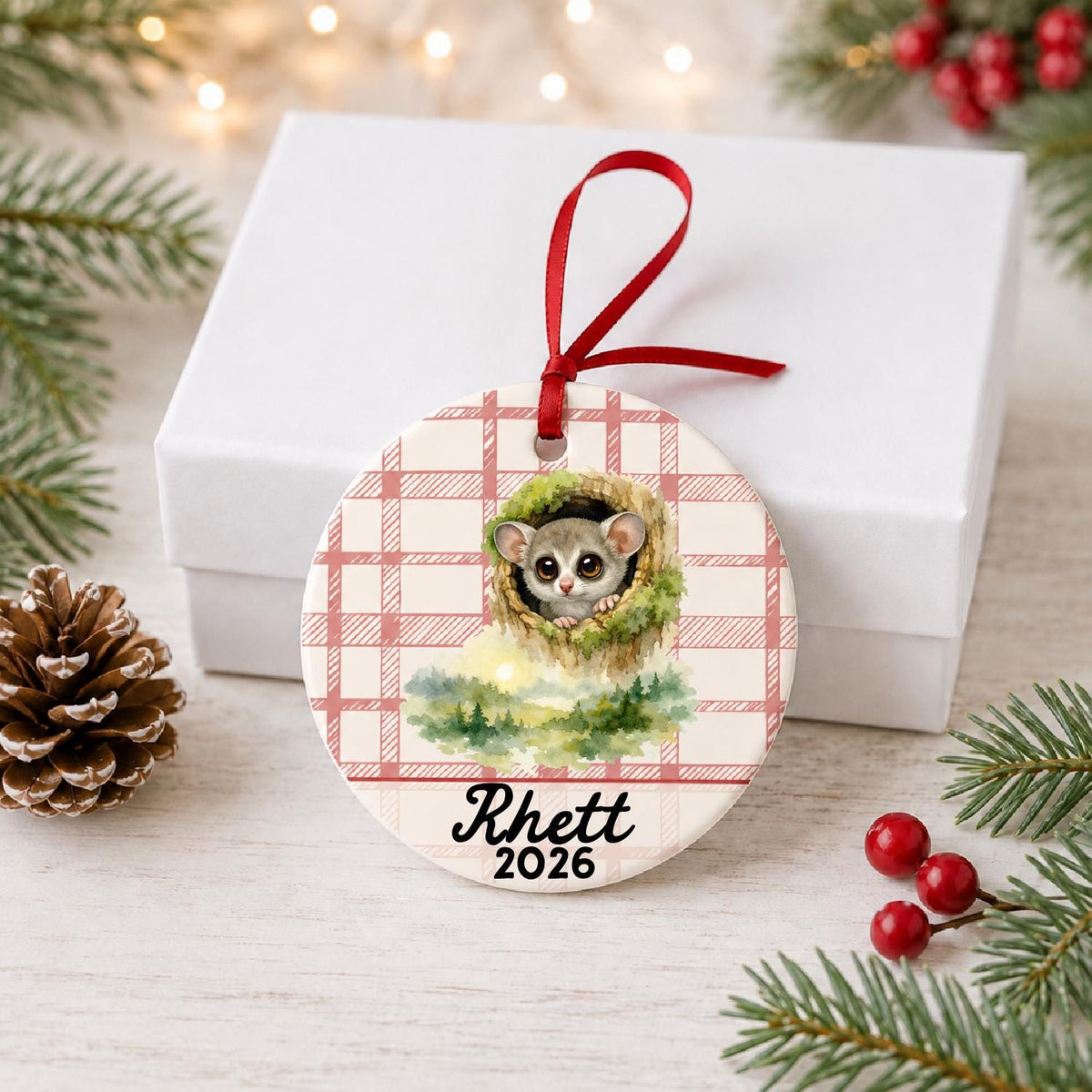 Personalized galago ceramic ornament with name and year on red plaid in front of a white gift box with pinecones and red berries