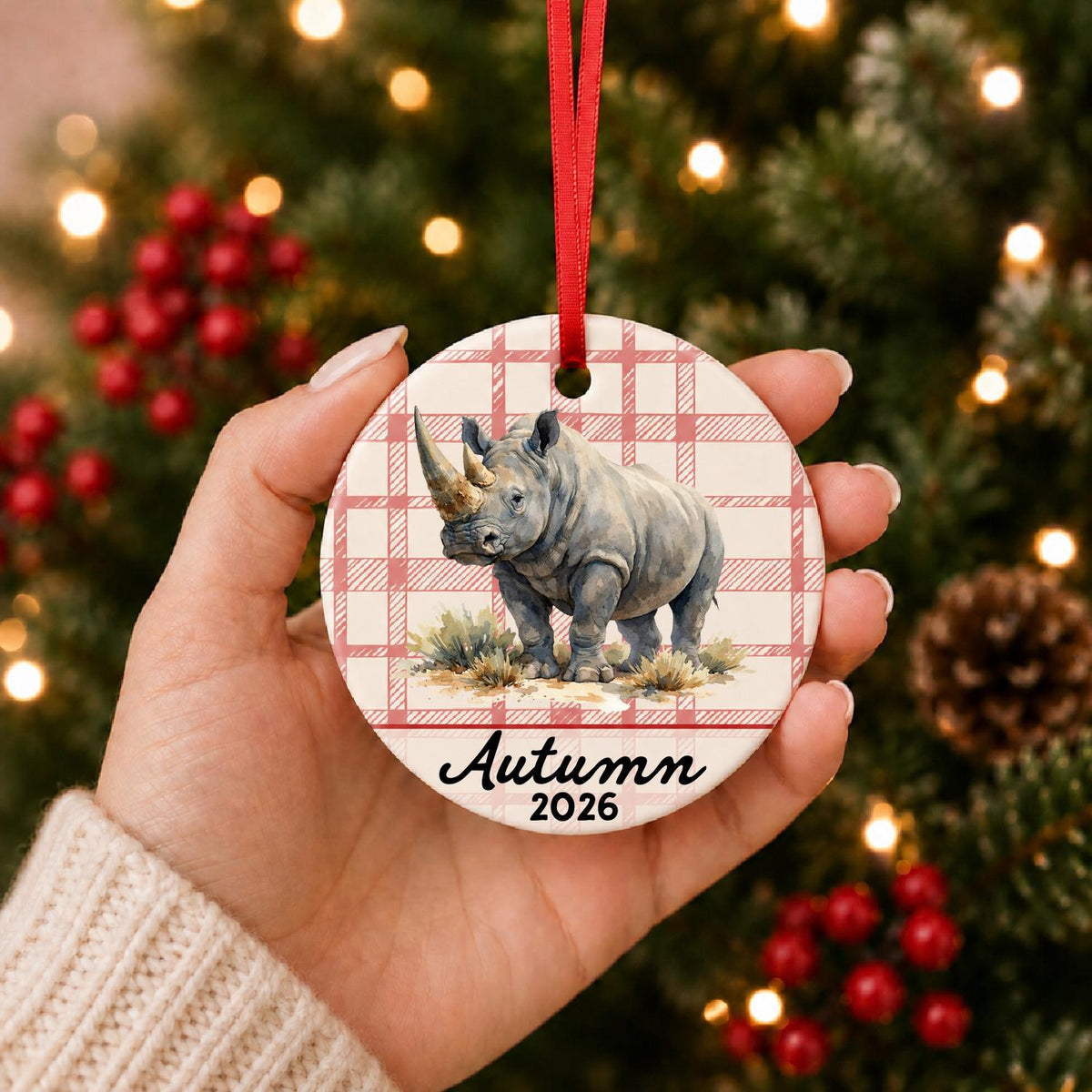 Hand holding the personalized white rhinoceros in grassland ceramic ornament with name and year on red plaid in front of a Christmas tree
