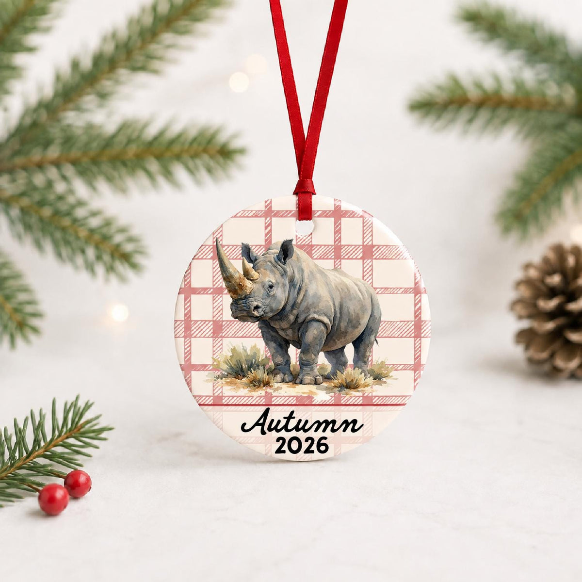 Personalized white rhinoceros in grassland ceramic ornament with name and year on red plaid with red ribbon on a white surface with pine branches and pinecones