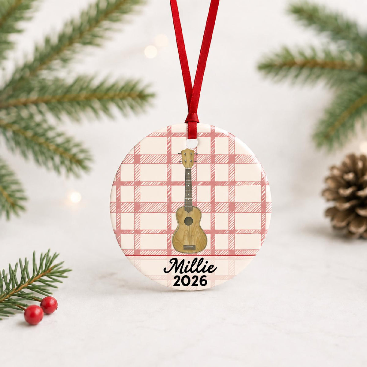 Personalized ukulele ceramic ornament with name and year on red plaid with red ribbon on a white surface with pine branches and pinecones
