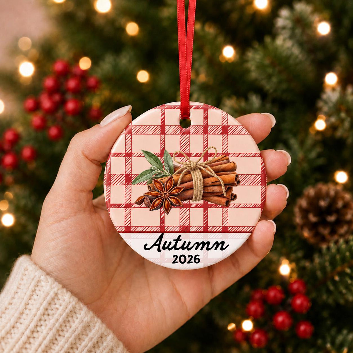 Hand holding the personalized cinnamon stick ceramic ornament with name and year on red plaid in front of a Christmas tree