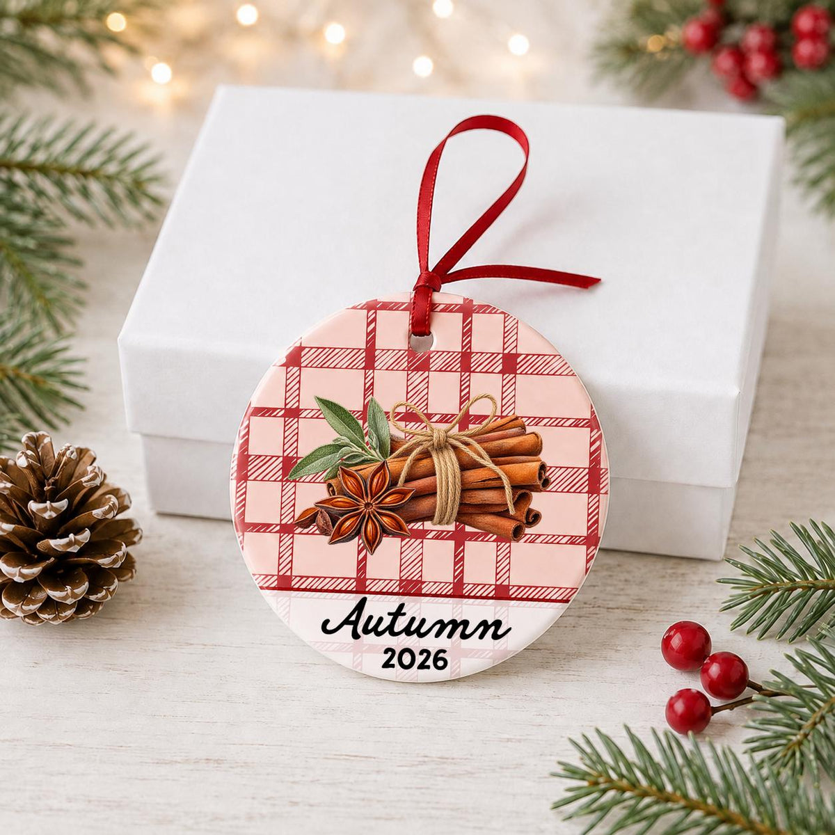 Personalized cinnamon stick ceramic ornament with name and year on red plaid in front of a white gift box with pinecones and red berries