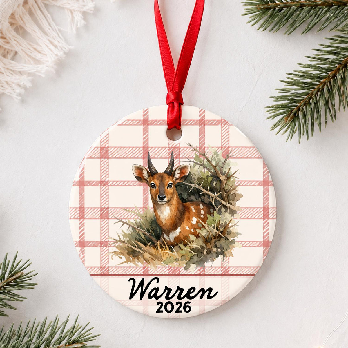 Personalized bushbuck ceramic ornament with name and year on red plaid styled with pine branches on a soft white background
