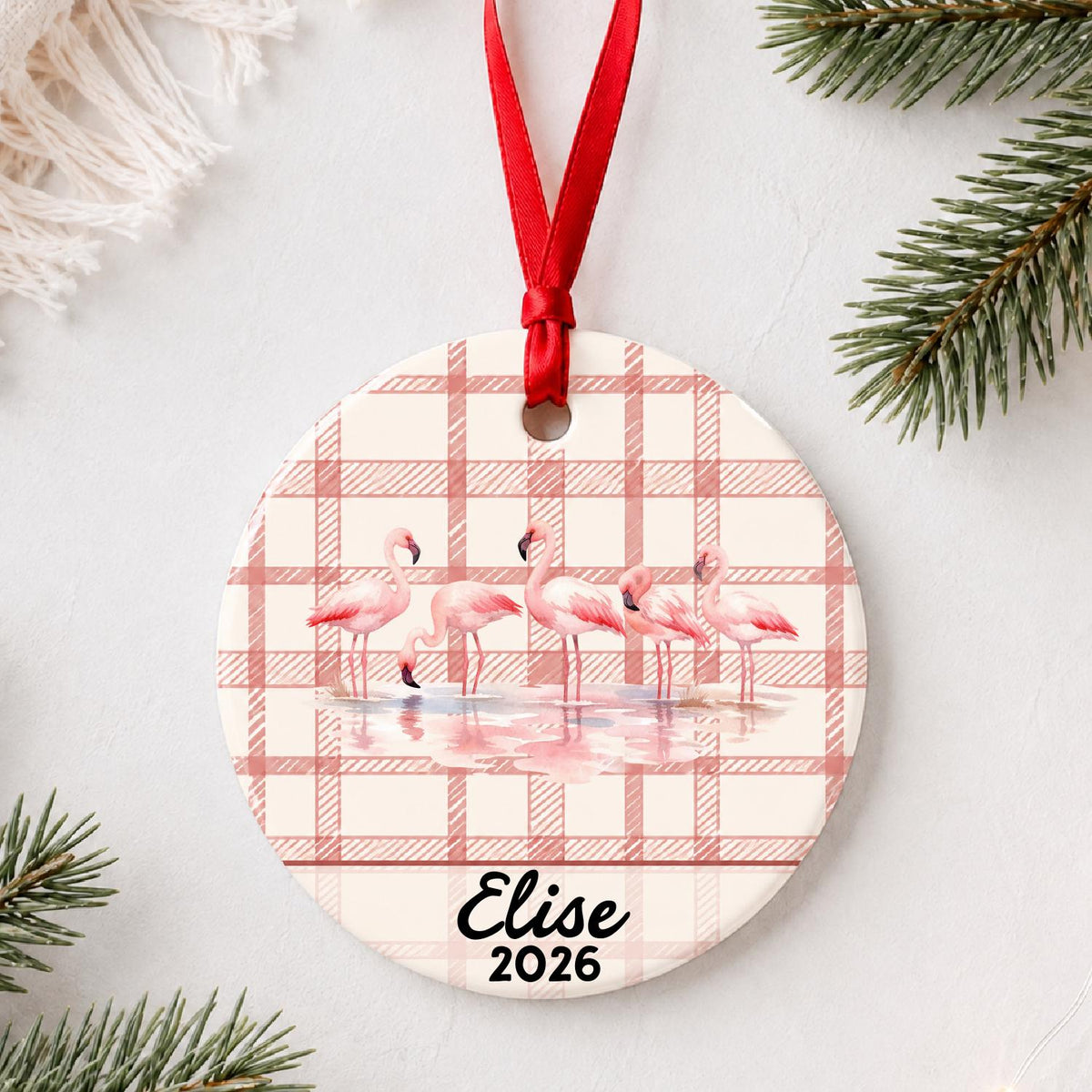 Personalized flamingo flock ceramic ornament with name and year on red plaid styled with pine branches on a soft white background