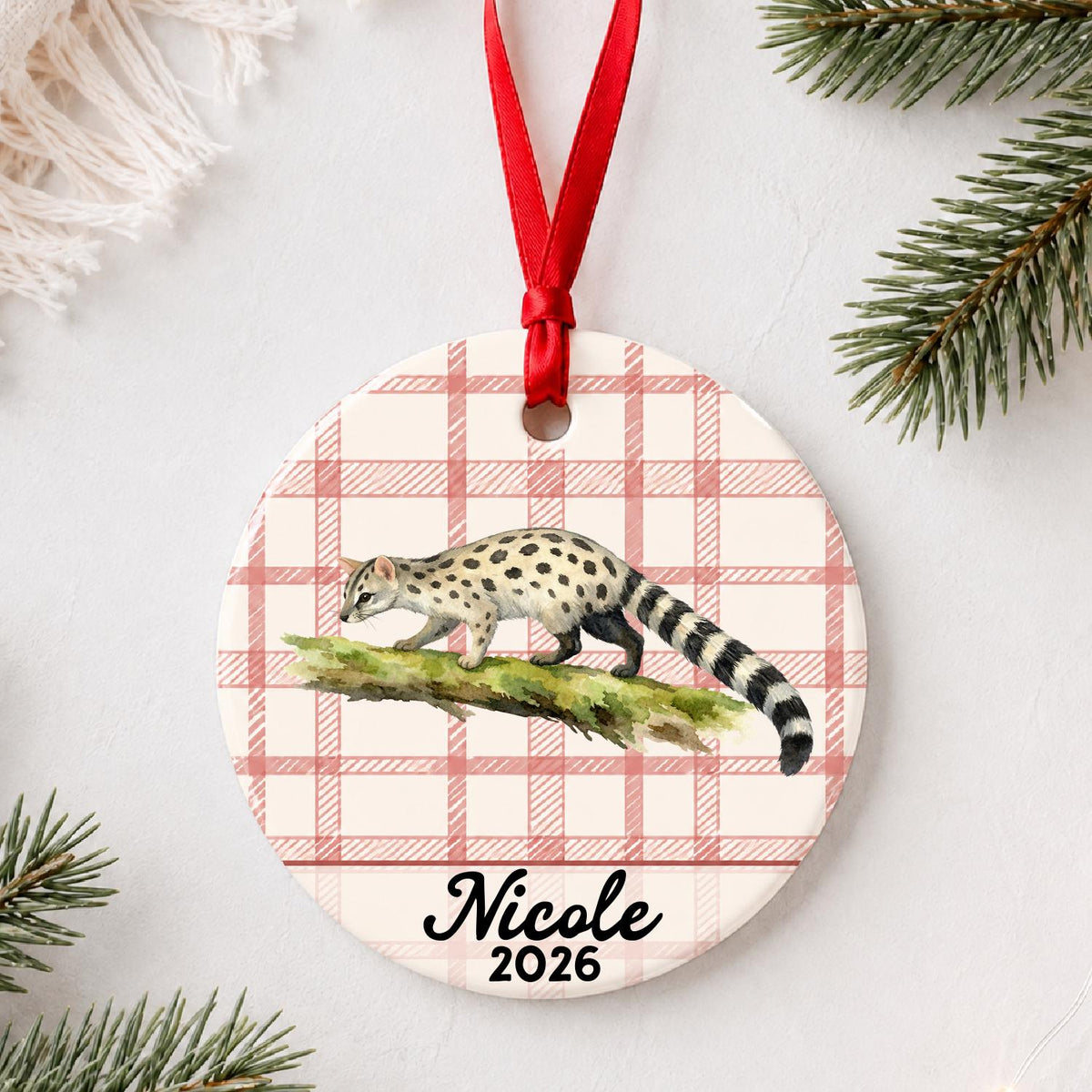 Personalized genet ceramic ornament with name and year on red plaid styled with pine branches on a soft white background