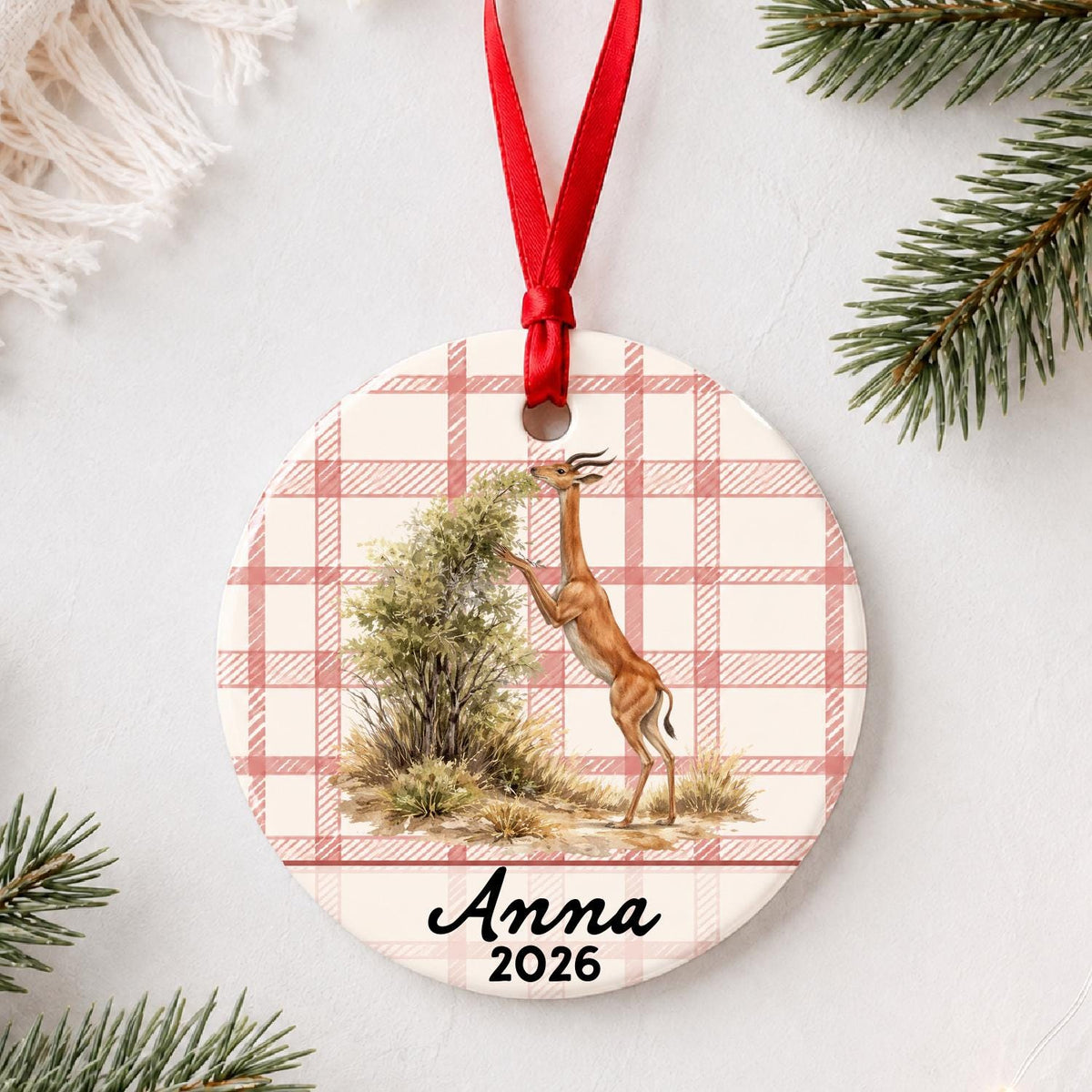 Personalized gerenuk ceramic ornament with name and year on red plaid styled with pine branches on a soft white background