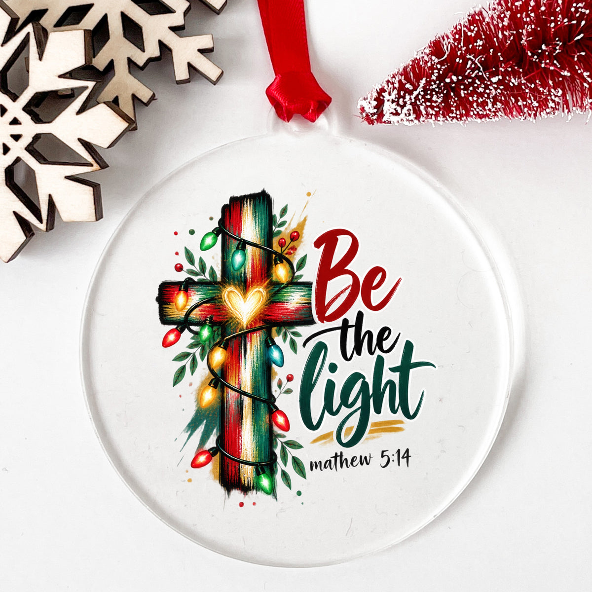 Be The Light Cross With Christmas Lights UV DTF sticker applied to a clear round acrylic ornament with a red ribbon