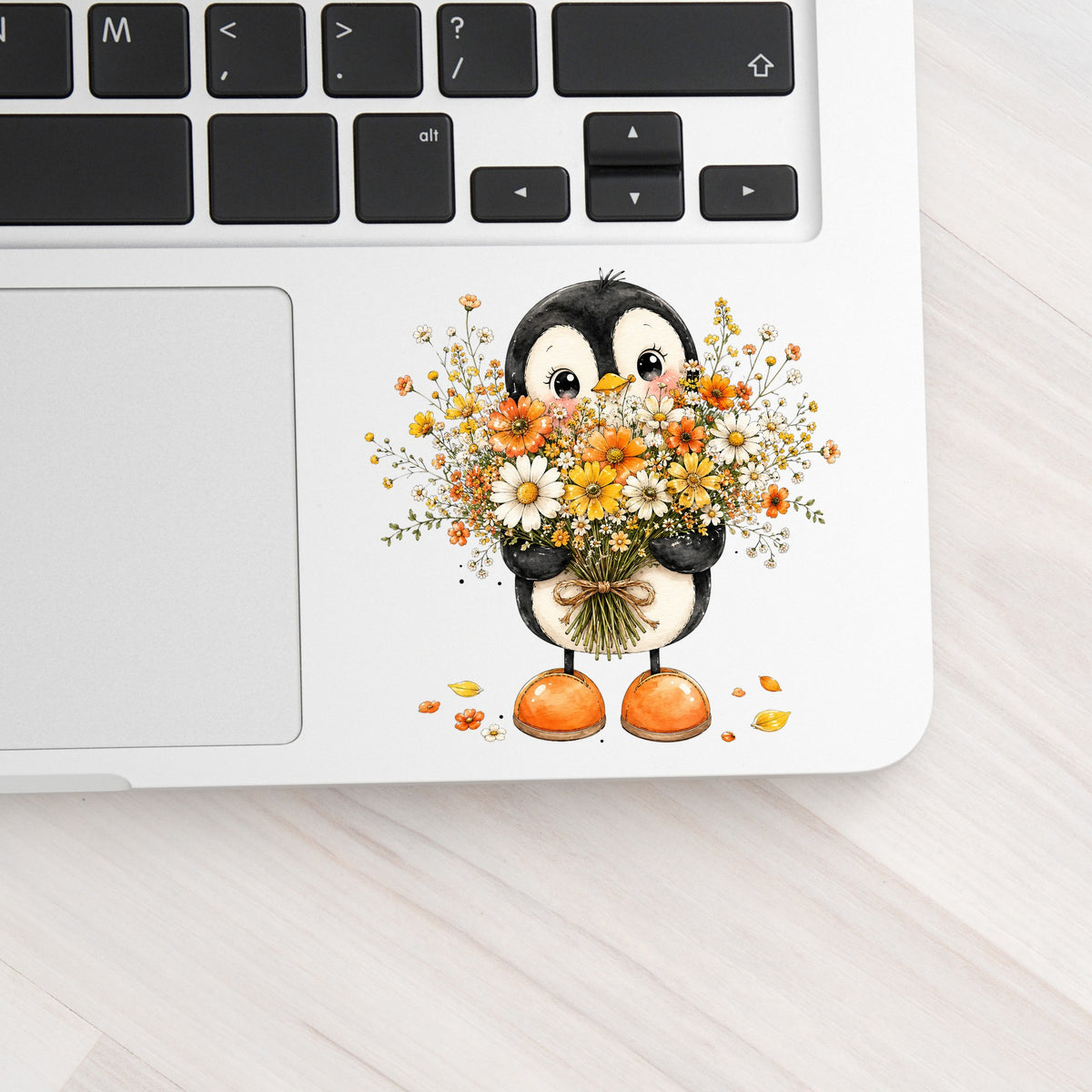 Baby Penguin Holding Wildflower Bouquet UV DTF sticker applied to a silver laptop beside the keyboard