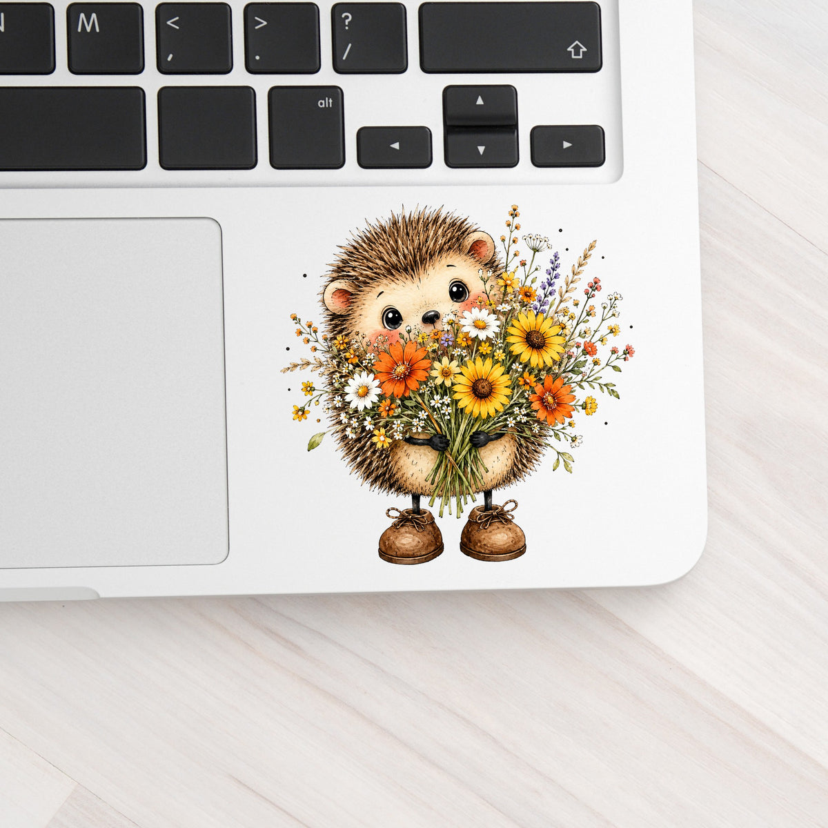 Hedgehog Holding Wildflower Bouquet UV DTF sticker applied to a silver laptop beside the keyboard