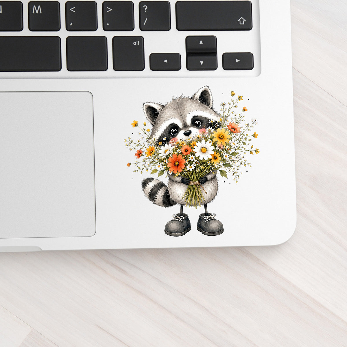 Raccoon Holding Wildflower Bouquet UV DTF sticker applied to a silver laptop beside the keyboard