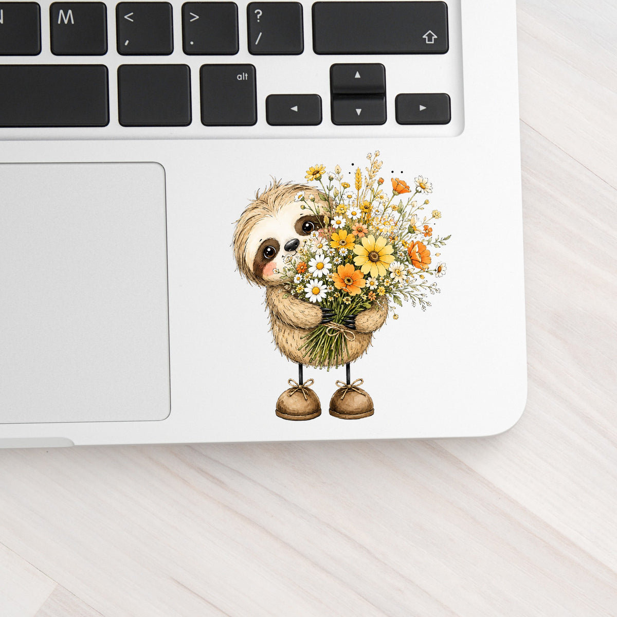 Baby Sloth Holding Wildflower Bouquet UV DTF sticker applied to a silver laptop beside the keyboard
