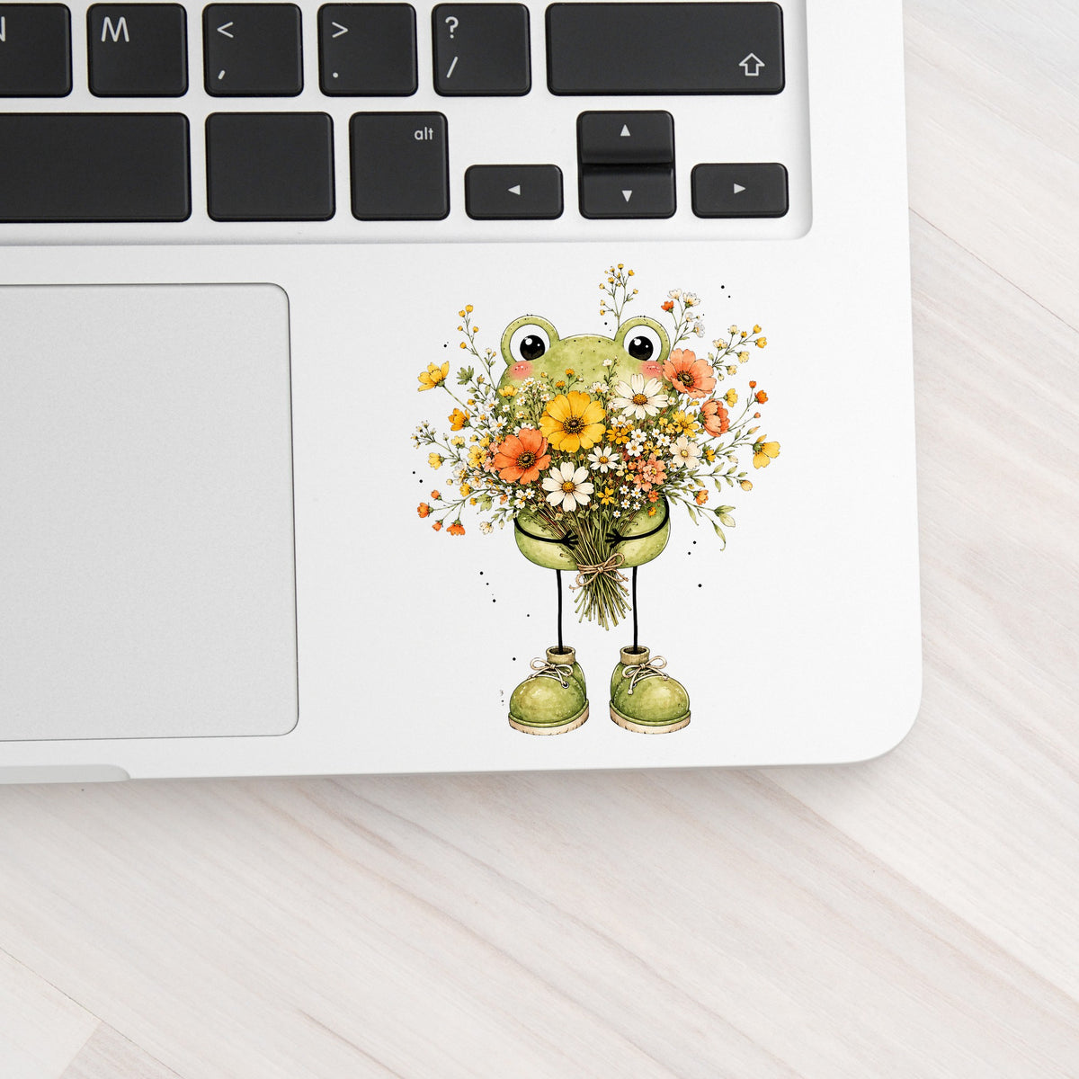 Green Frog Holding Wildflower Bouquet UV DTF sticker applied to a silver laptop beside the keyboard