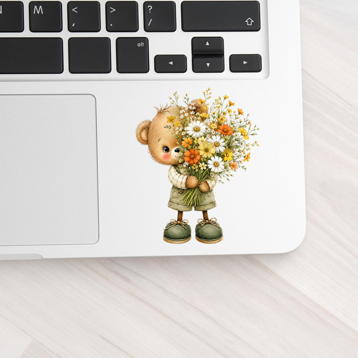 Brown Bear In Overalls Holding Daisy Bouquet UV DTF sticker applied to a silver laptop beside the keyboard