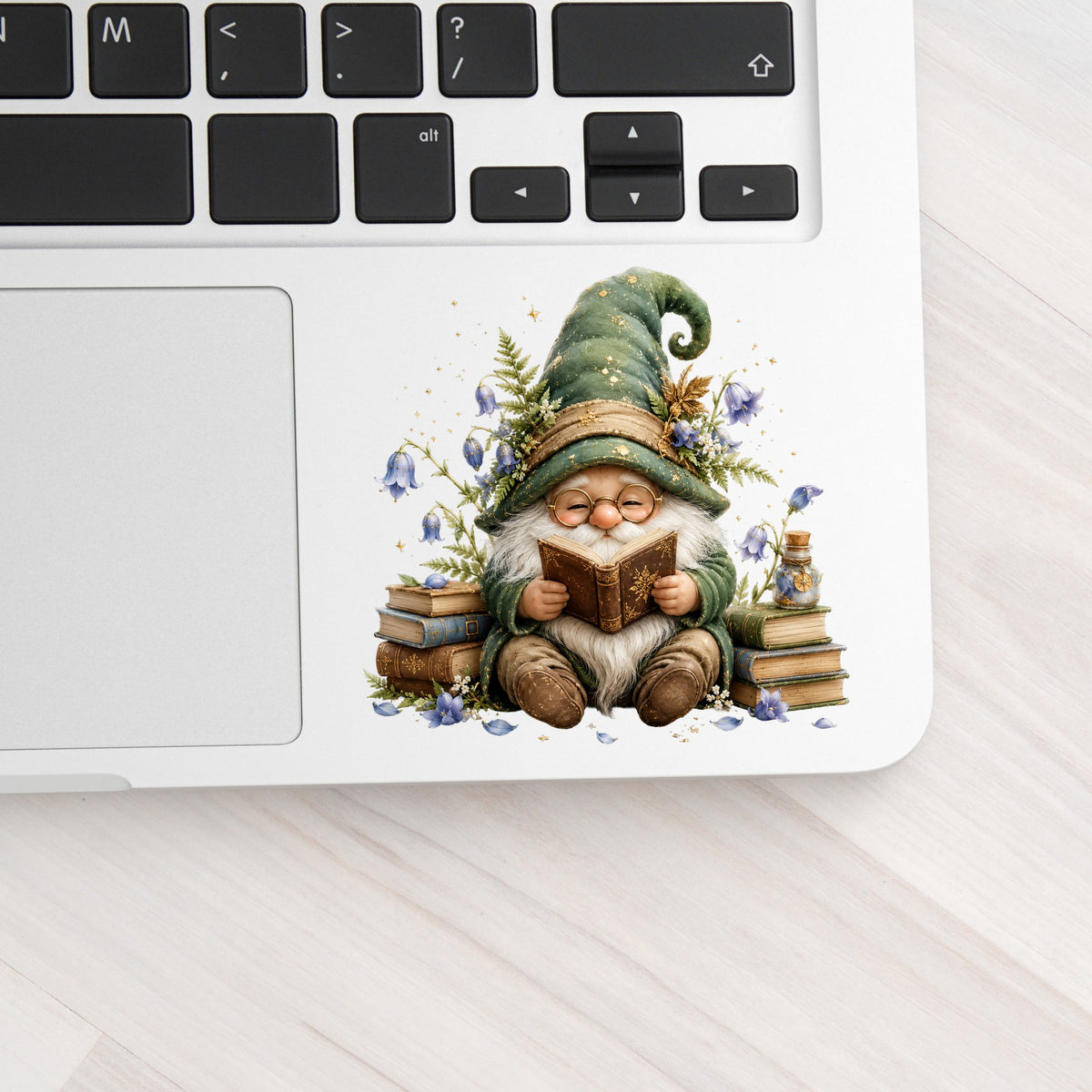 Gnome Reading With Book Stack UV DTF sticker applied to a silver laptop beside the keyboard