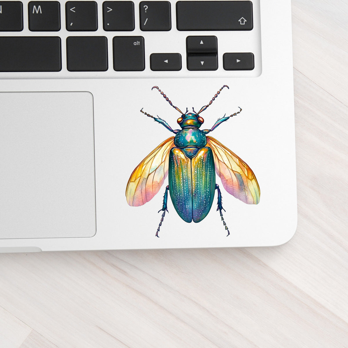 Golden Winged Flying Beetle UV DTF sticker applied to a silver laptop beside the keyboard