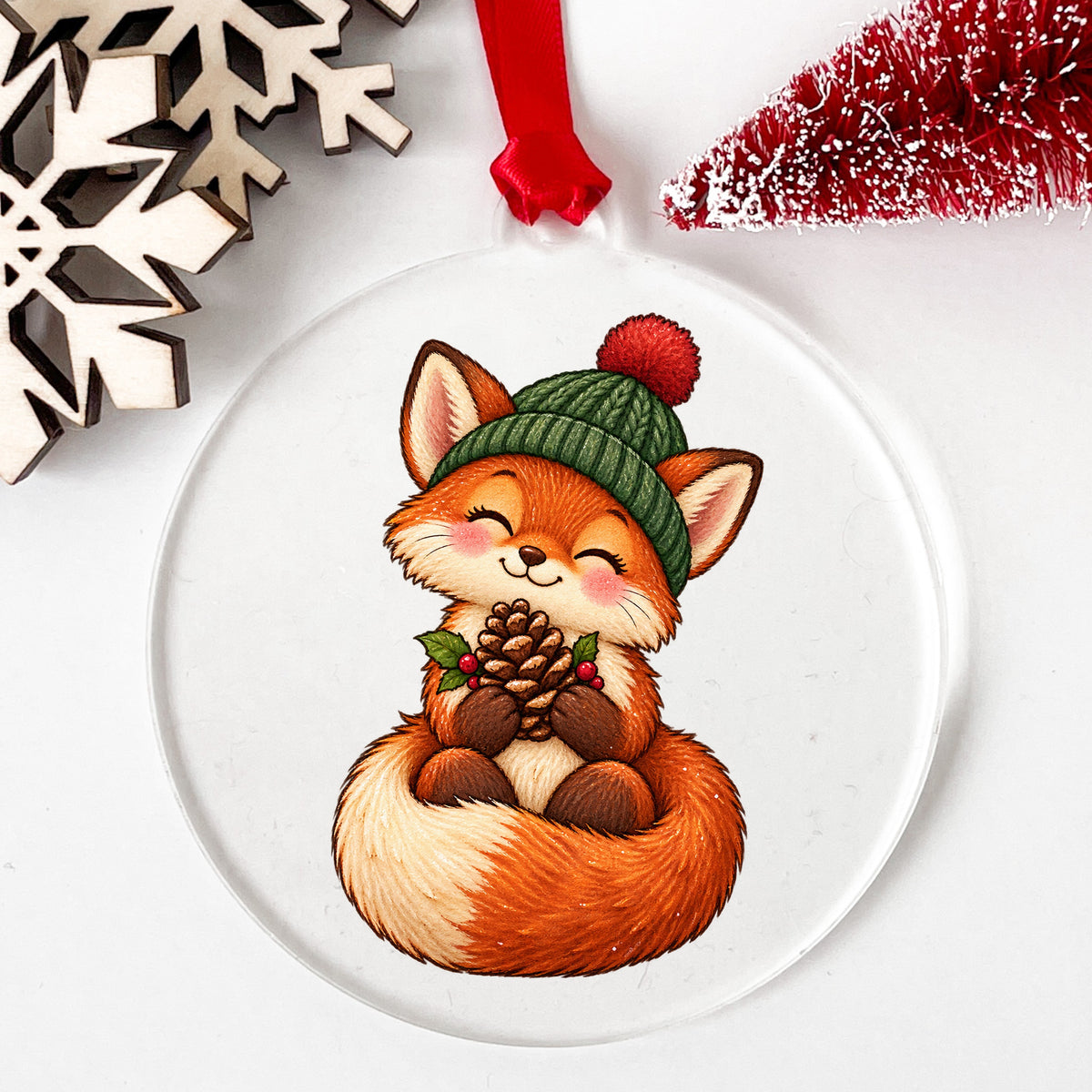 Red Fox In Knit Beanie Holding Pinecone UV DTF sticker applied to a clear round acrylic ornament with a red ribbon