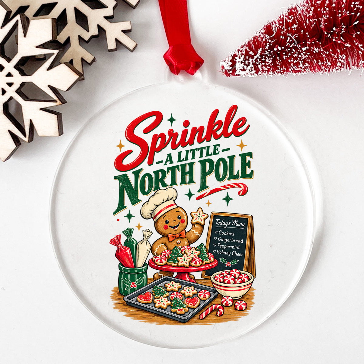 Sprinkle A Little North Pole Gingerbread UV DTF sticker applied to a clear round acrylic ornament with a red ribbon